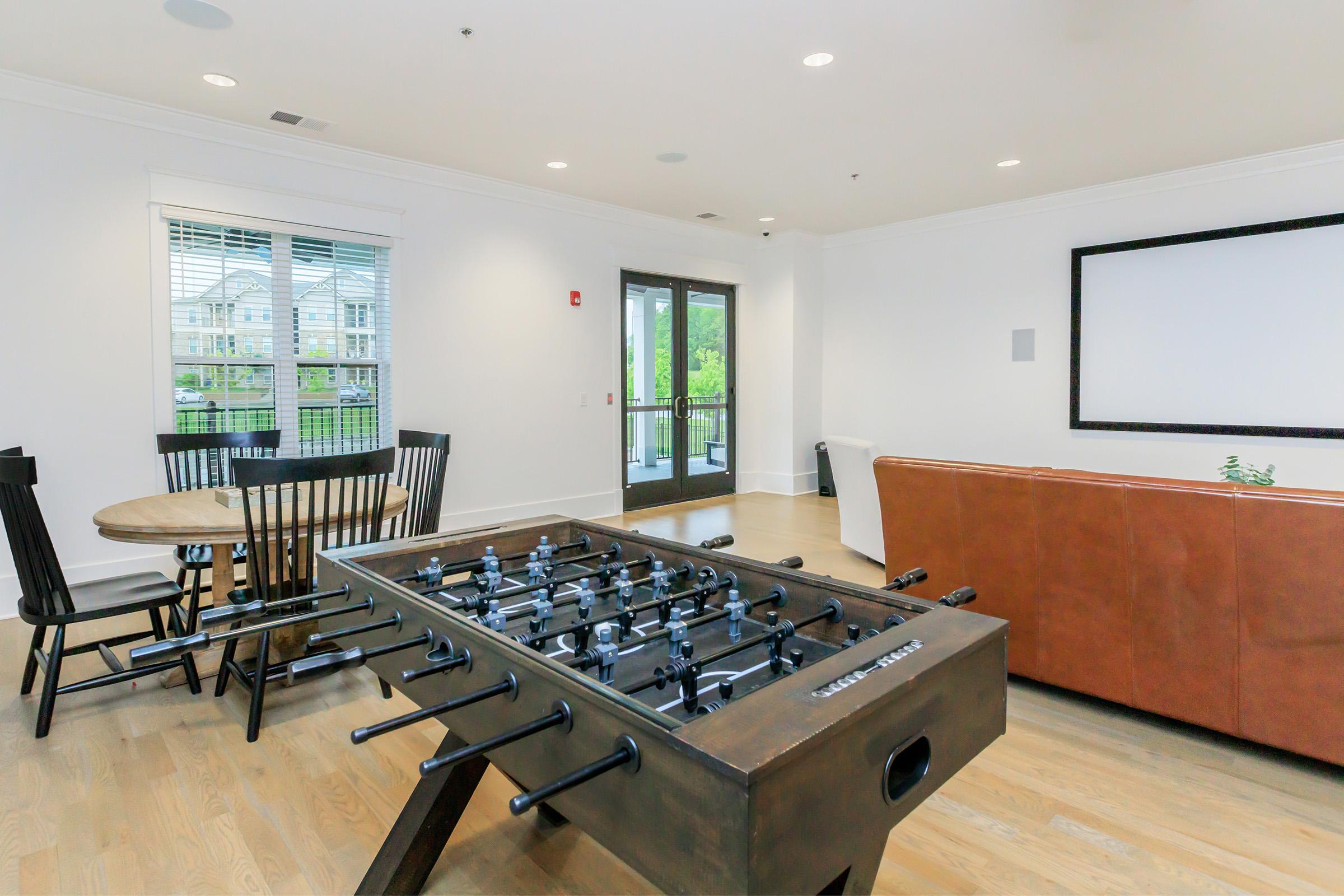 A modern living space featuring a foosball table in the foreground, a dining table with chairs to the left, a comfortable brown couch, and large windows letting in natural light. The walls are painted white, creating a bright and airy atmosphere. A sliding door leads to an outdoor area.