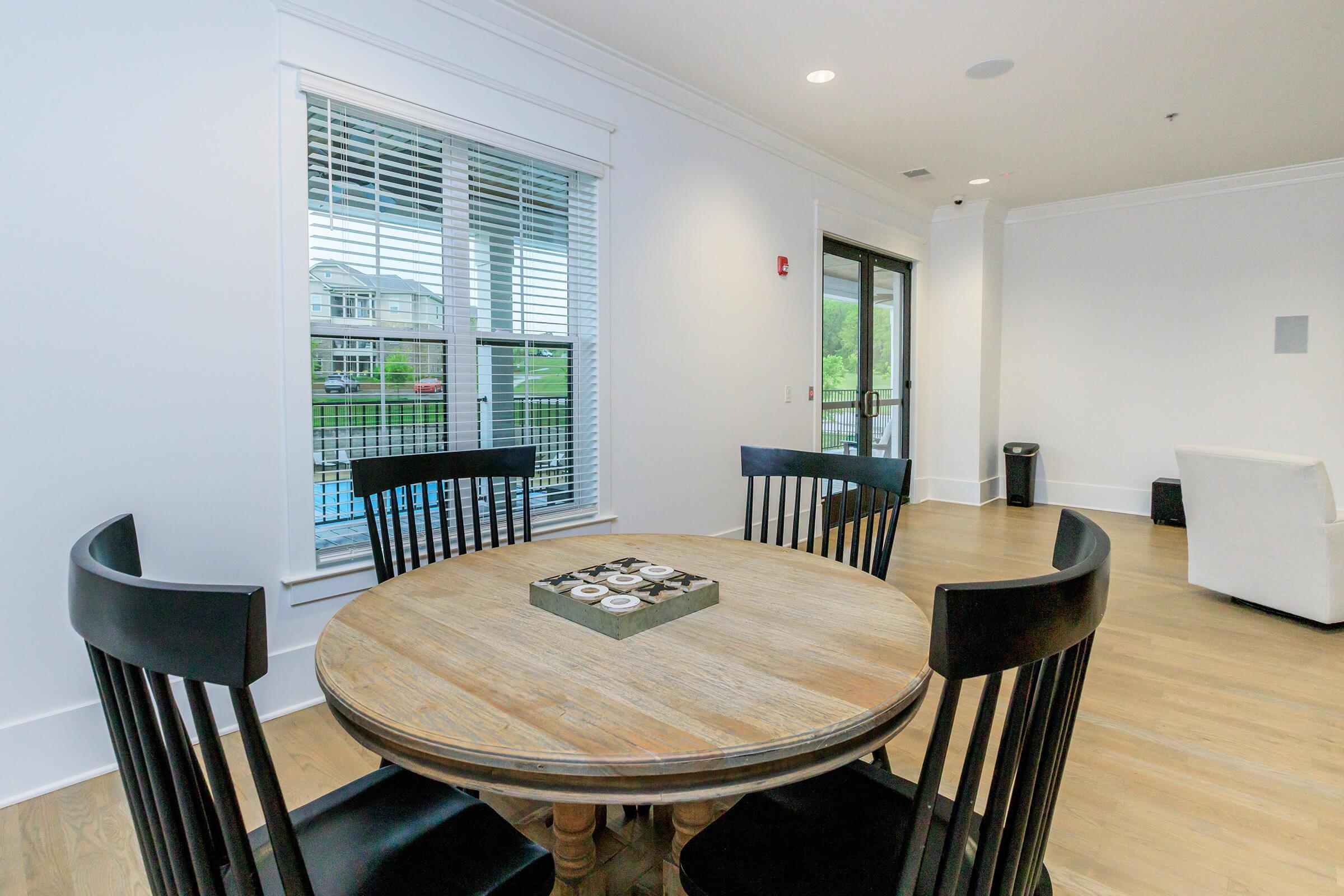 A round wooden table with four black chairs sits in a brightly lit room. A game board is centered on the table. Large windows provide a view of the outdoors, and a white couch is visible in the background. The walls are painted white, and the floor is made of light-colored wood.