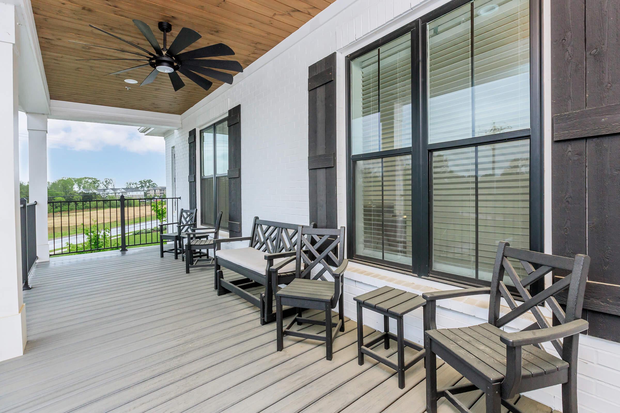 A spacious porch with wooden flooring, featuring a ceiling fan and large, divided windows. There are two sets of black outdoor chairs and a bench positioned against the white walls, adding a cozy and inviting atmosphere. The backdrop includes a view of greenery and open fields.