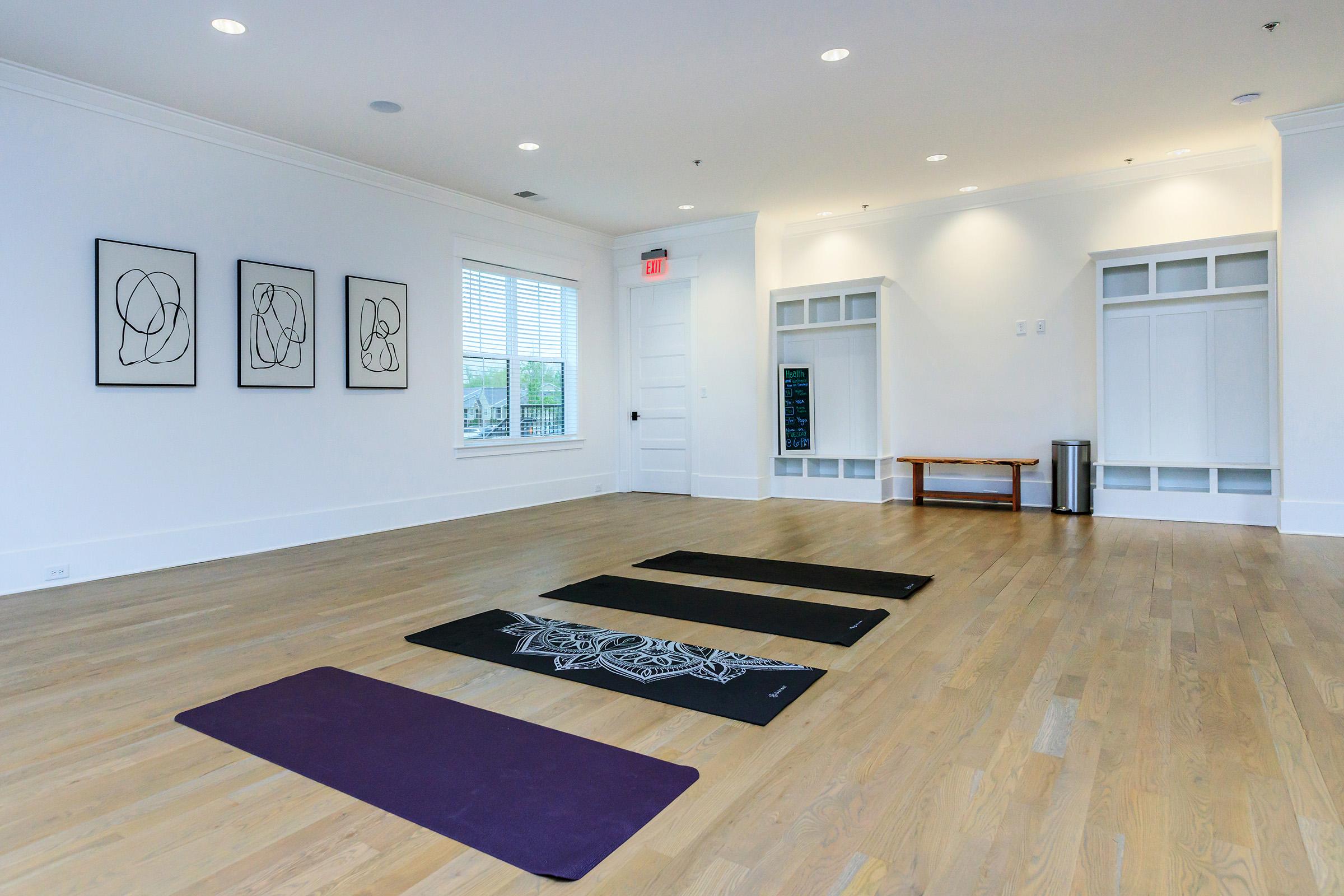 A clean, spacious yoga studio featuring three yoga mats on a light wooden floor. The walls are adorned with minimalist art pieces, and large windows allow natural light to fill the room. There's a bench against the wall and a small trash can nearby, creating a calm and inviting atmosphere for practice.