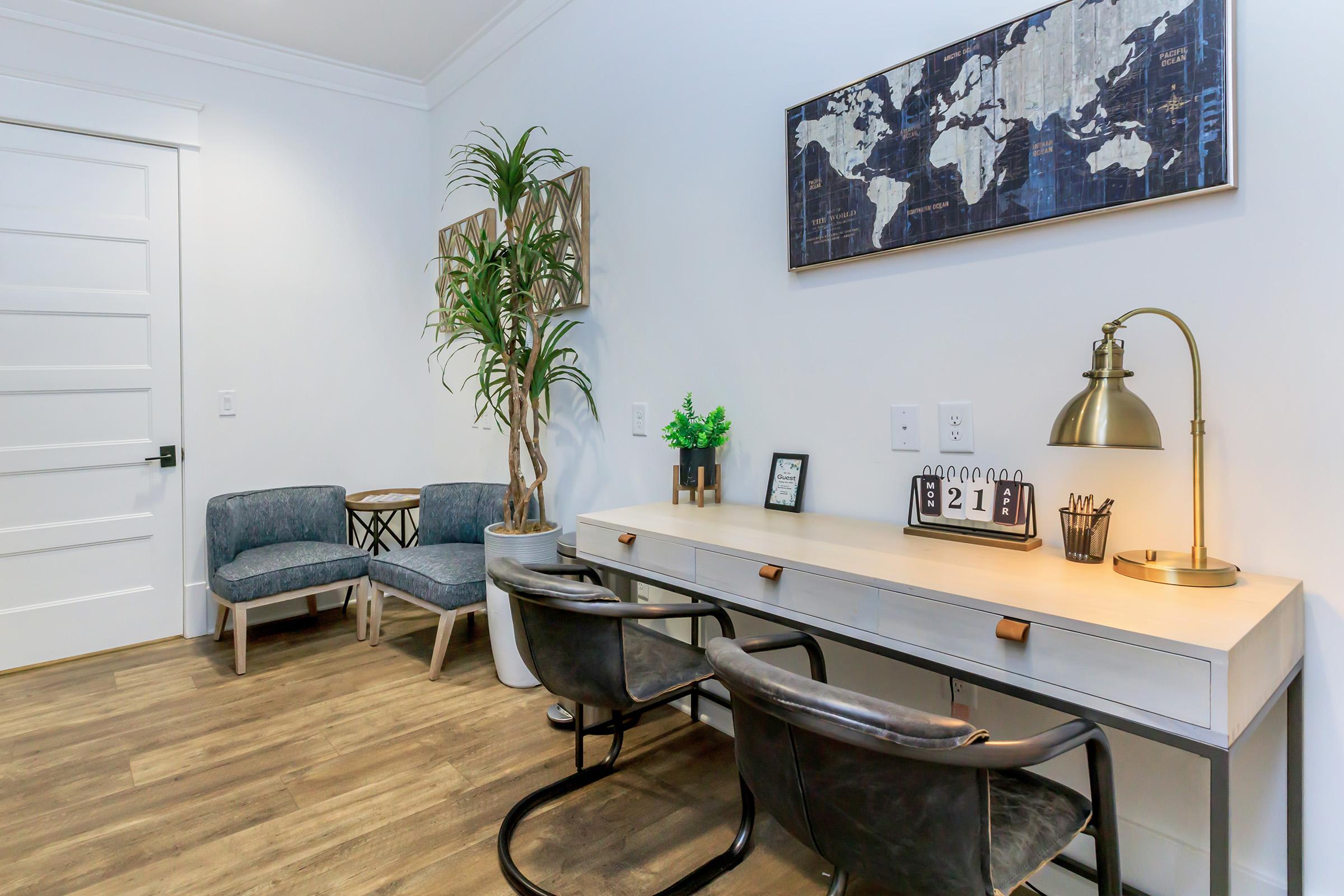 A modern home office with a light wooden desk and two black metal chairs. There are decorative plants, a wall art map, and a small calendar on the desk. In the background, two blue upholstered armchairs and a round side table create a cozy seating area. The walls are painted white, enhancing the bright, airy atmosphere.
