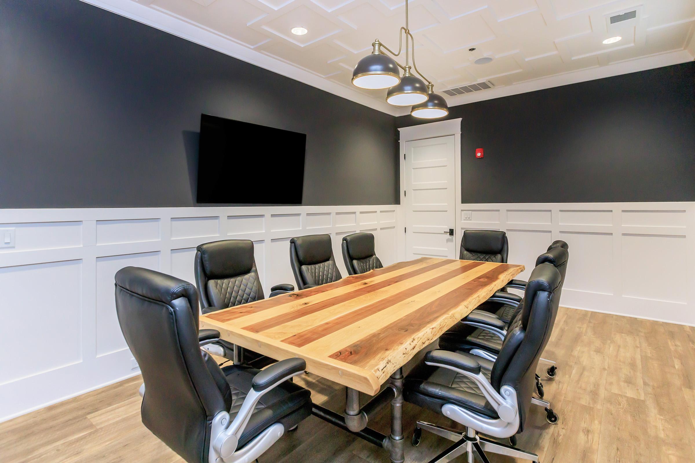 A modern conference room featuring a large wooden table surrounded by six black ergonomic chairs. The room has dark grey walls with white wainscoting and includes a flat-screen TV mounted on the wall. The decor is minimalist with overhead lighting fixtures and a wooden floor.