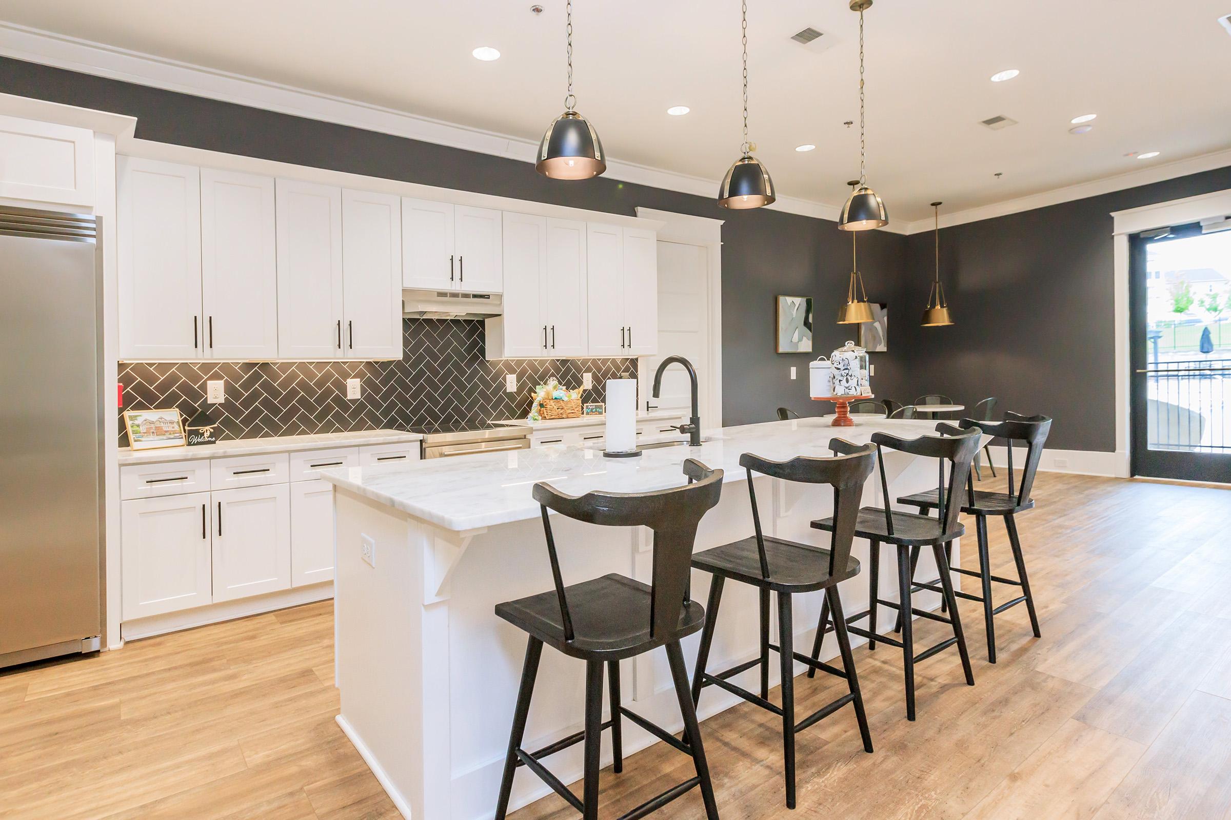 A modern kitchen featuring a long island with black wooden stools, white cabinetry, and a marble countertop. The backsplash is a dark herringbone pattern, with pendant lights hanging above. Large windows provide natural light, enhancing the contemporary design.