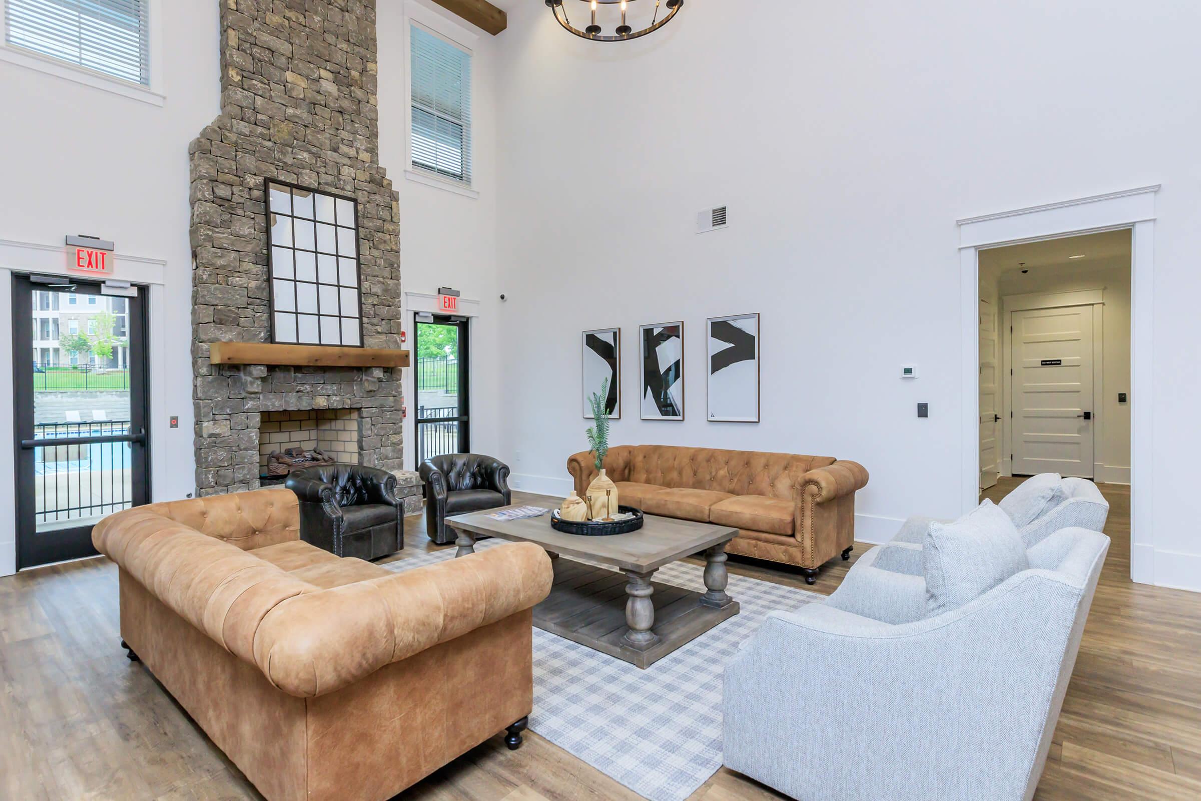 A spacious living room with a large stone fireplace, featuring two brown leather sofas, a light gray sofa, and a wooden coffee table. The room has large windows with natural light, and modern wall art is displayed. Light hardwood flooring adds a warm touch to the contemporary design.