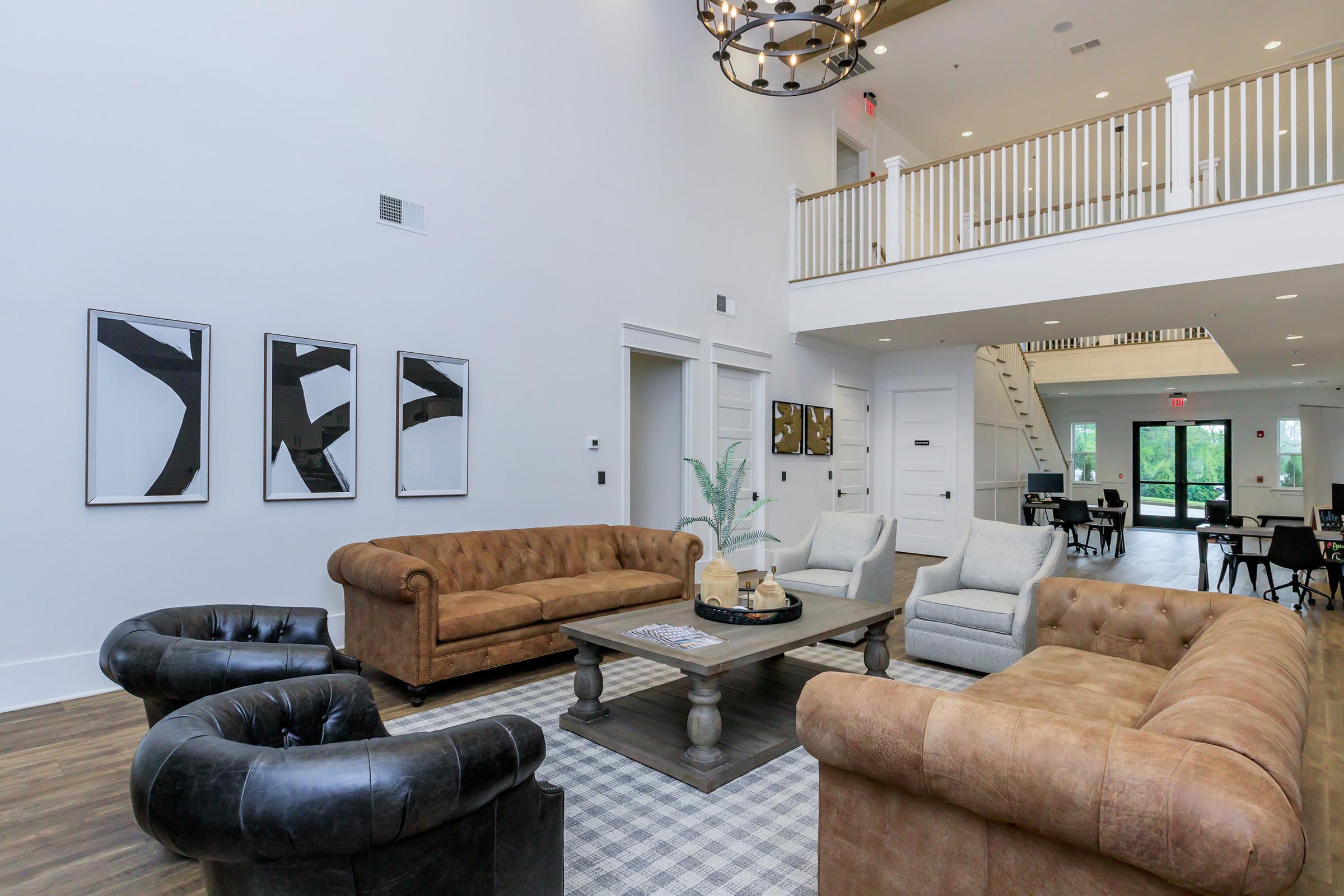 A spacious lobby with high ceilings featuring two chesterfield sofas in brown and black leather. There are two modern chairs, a wooden coffee table with decorative items, and abstract wall art. The area is well-lit with natural light coming through large windows and has a stylish chandelier.