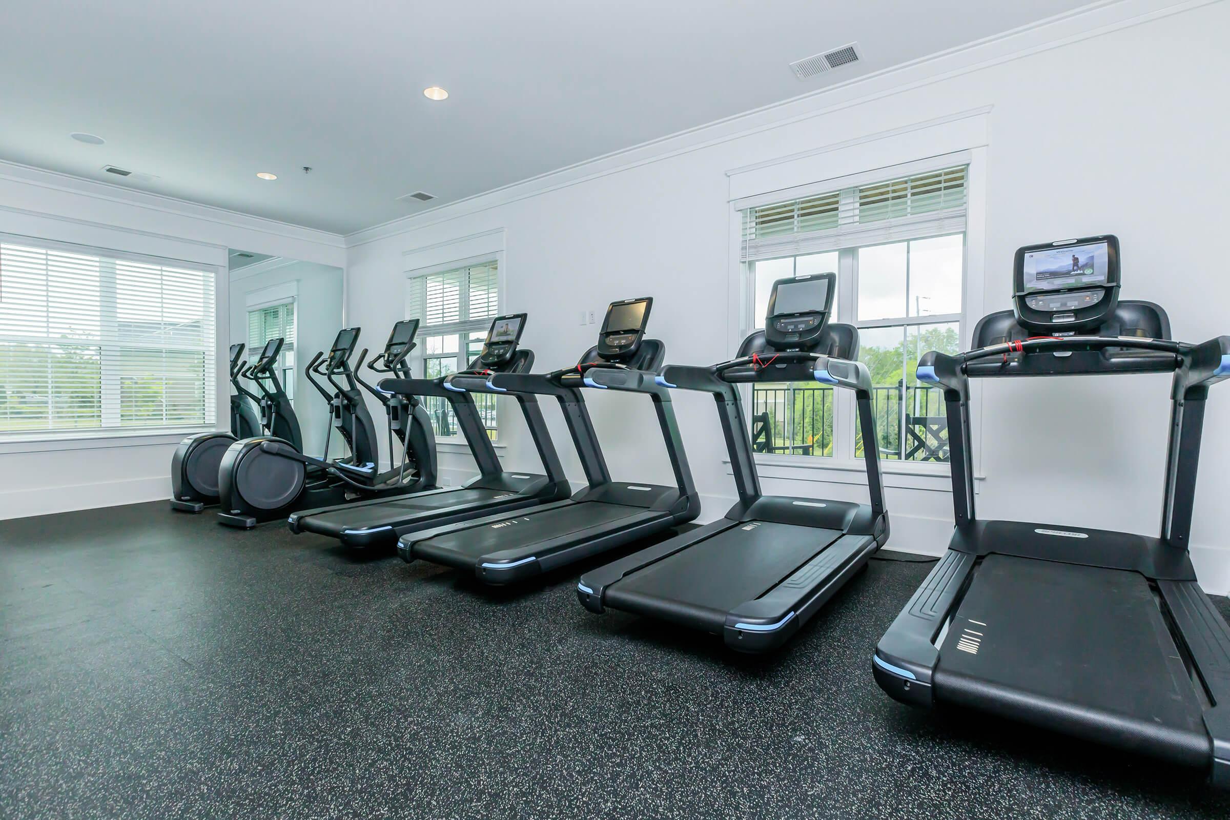 A modern gym interior featuring multiple treadmills lined up against a wall. Large windows allow natural light to fill the space, with a view of greenery outside. The room has a clean, bright aesthetic with a rubber floor for safety and exercise.