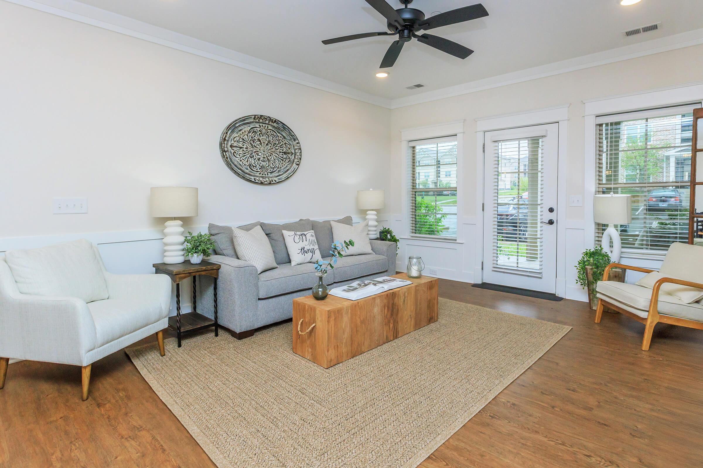 A modern living room featuring a light gray sofa with decorative pillows, a wooden coffee table, and two armchairs. Natural light streams in through windows with blinds. There are two lamps on side tables, and a circular wall decor piece adds an artistic touch. A woven rug complements the warm wooden floors.