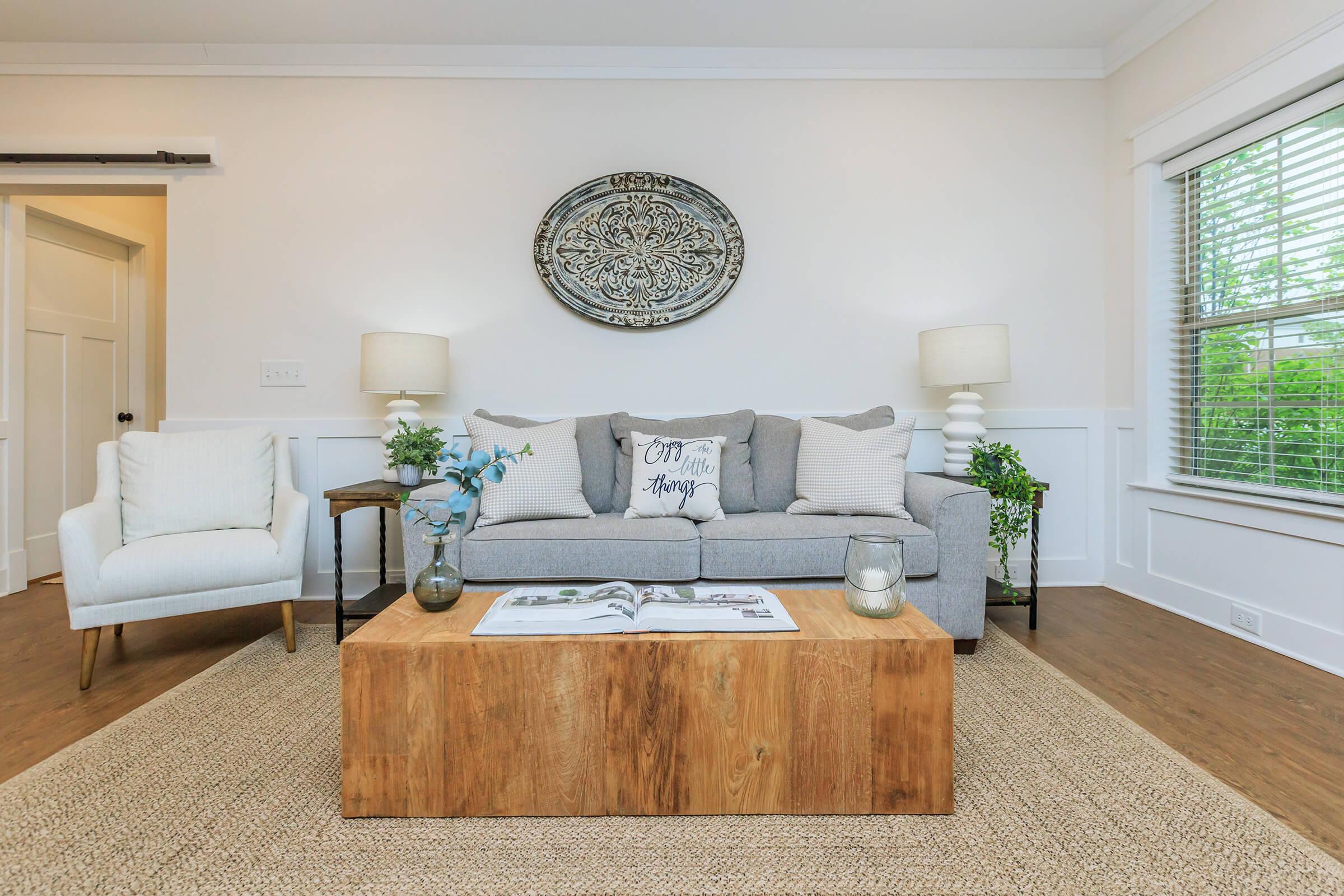 A cozy living room featuring a gray sofa with decorative cushions, two lamps on either side, a wooden coffee table with a magazine, and a light-colored area rug. The walls are adorned with a circular decorative element, and there's a window providing natural light.
