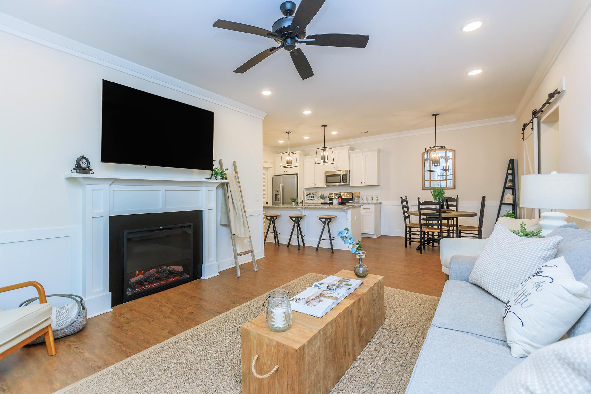 A modern living room featuring a gray sofa, a wooden coffee table, and a stylish ladder against the wall. There's a cozy fireplace beneath a large flat-screen TV, and a dining area with a table and chairs in the background. The space has a warm, inviting atmosphere with neutral colors and elegant decor.