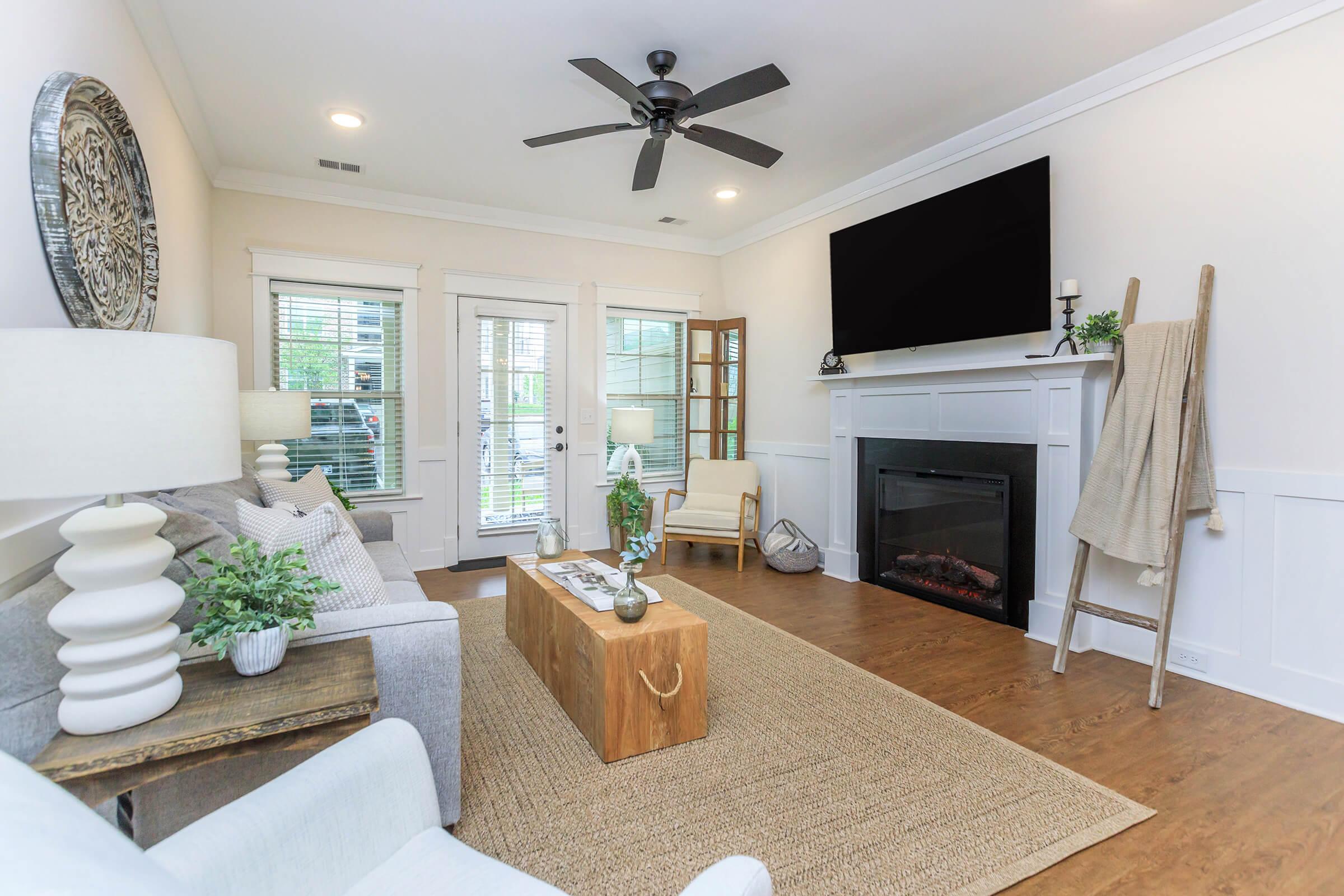 A cozy living room featuring a light-colored sofa and armchair, a wooden coffee table, and a plush rug. The space includes a fireplace with a modern TV above it, large windows with natural light, and a decorative ladder displaying a blanket. Green plants and stylish lamps add a welcoming touch.