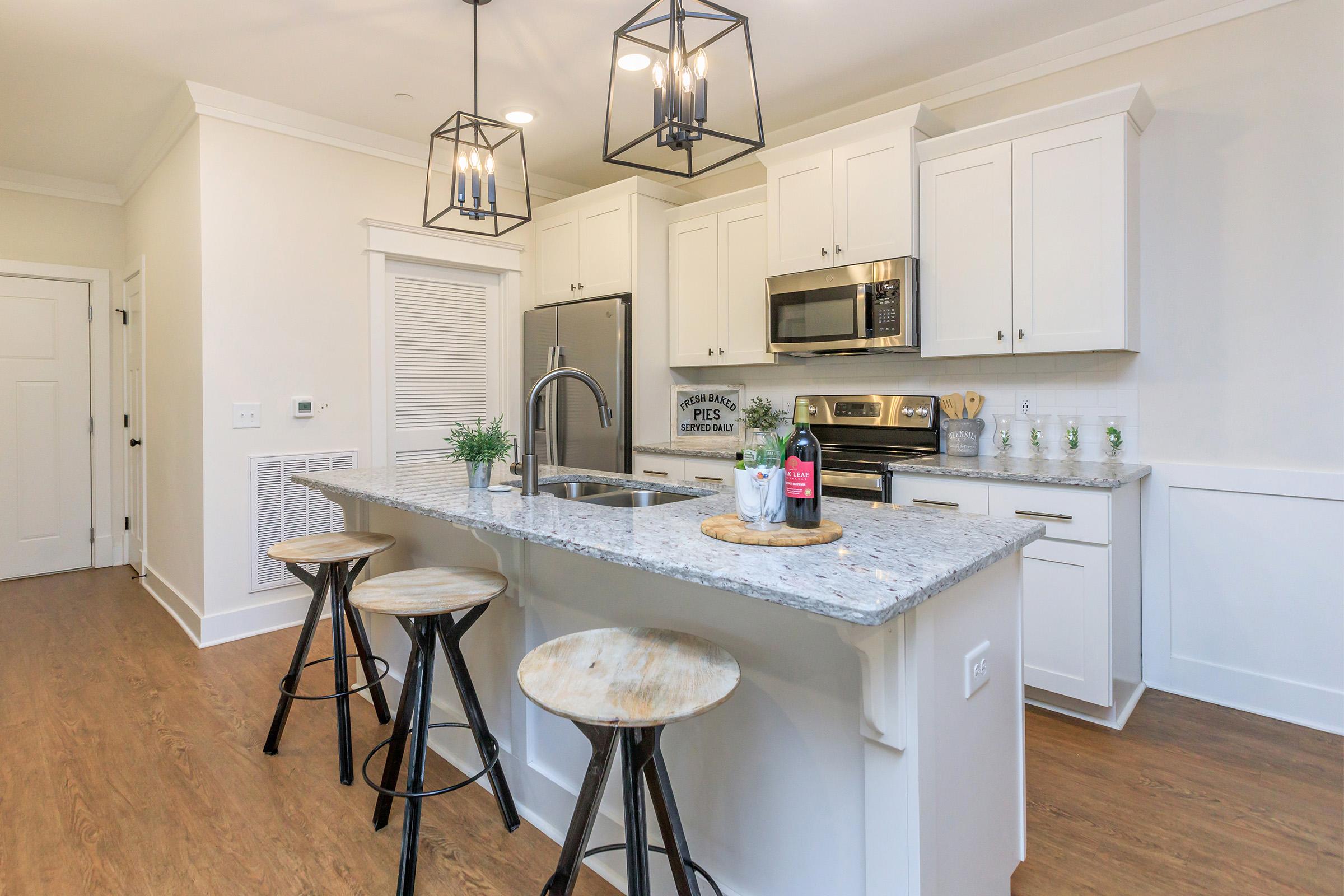 Modern kitchen featuring white cabinets, stainless steel appliances, and a large granite island with three wooden stools. Pendant lighting hangs above the island, and a small plant decorates the counter. The space has light-colored walls and hardwood flooring, creating a bright and inviting atmosphere.