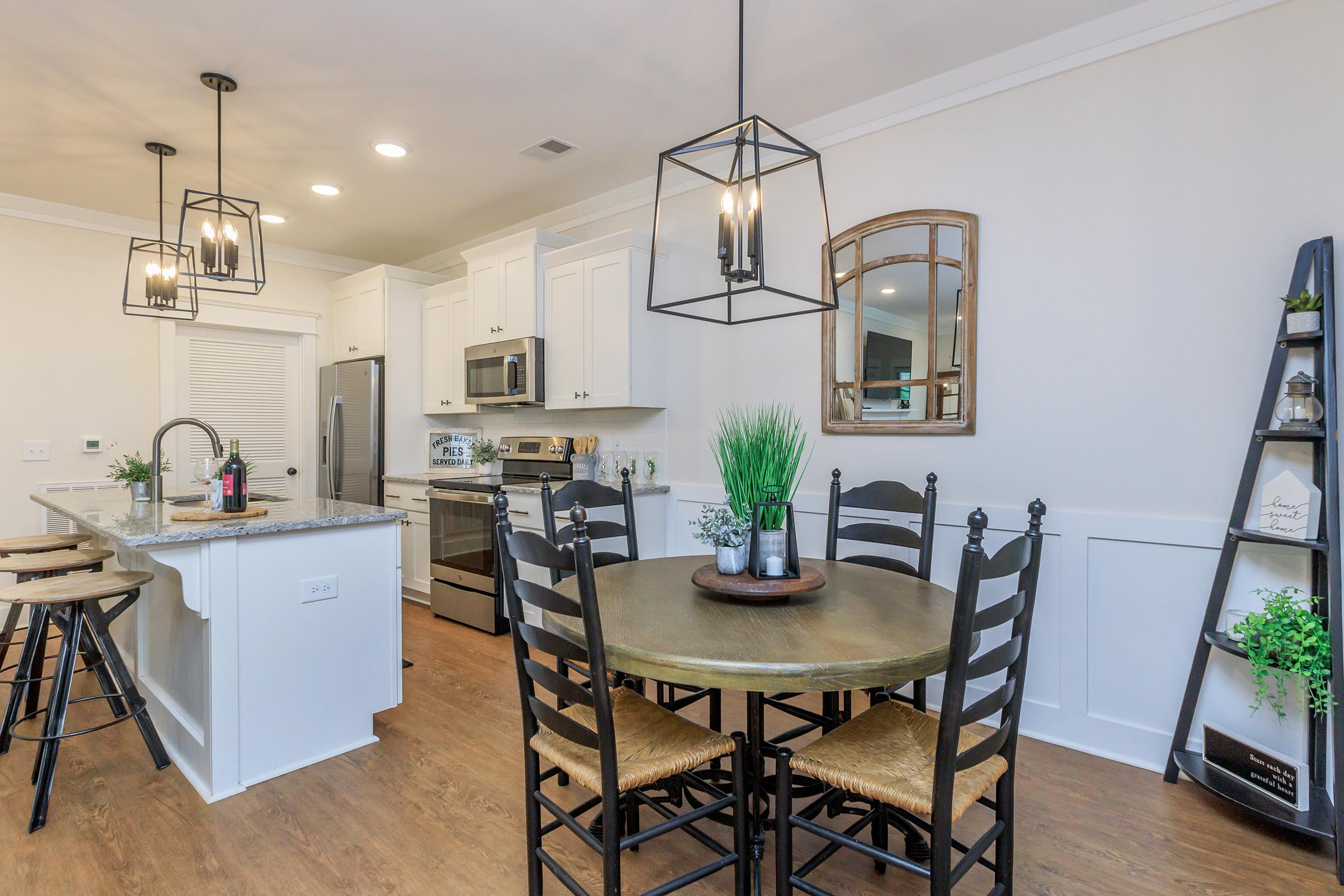A bright and modern kitchen and dining area featuring a round wooden table with black chairs, a stylish pendant light above, and a kitchen island with bar stools. The kitchen has white cabinetry, stainless steel appliances, and decorative plants. A mirror and small shelf are also part of the decor.