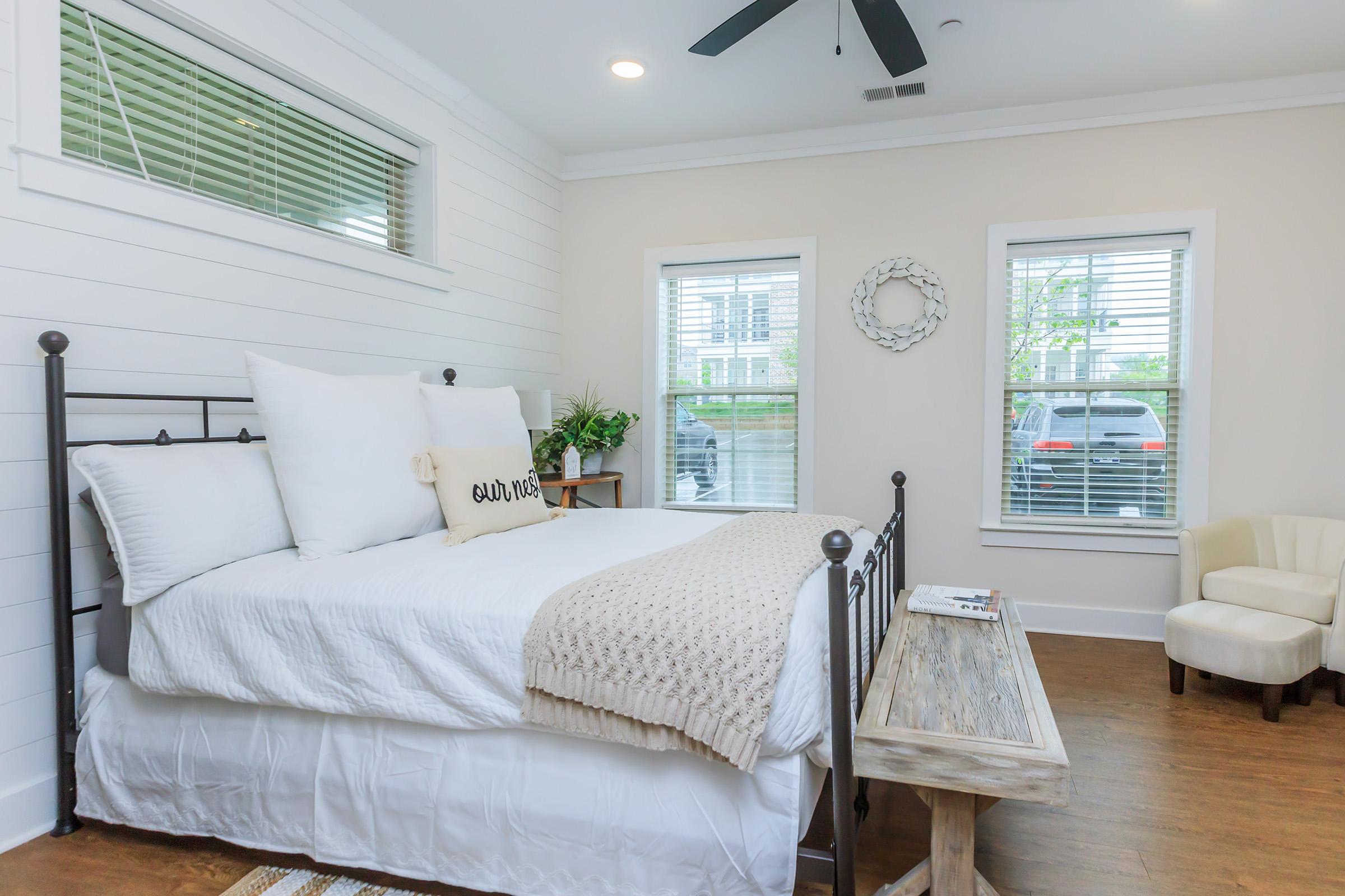 A cozy bedroom featuring a black metal bed frame with white bedding, two windows with blinds, a small plant on a bedside table, and a beige armchair. A decorative wall hanging and a white throw blanket add warmth to the space. Natural light brightens the inviting atmosphere.