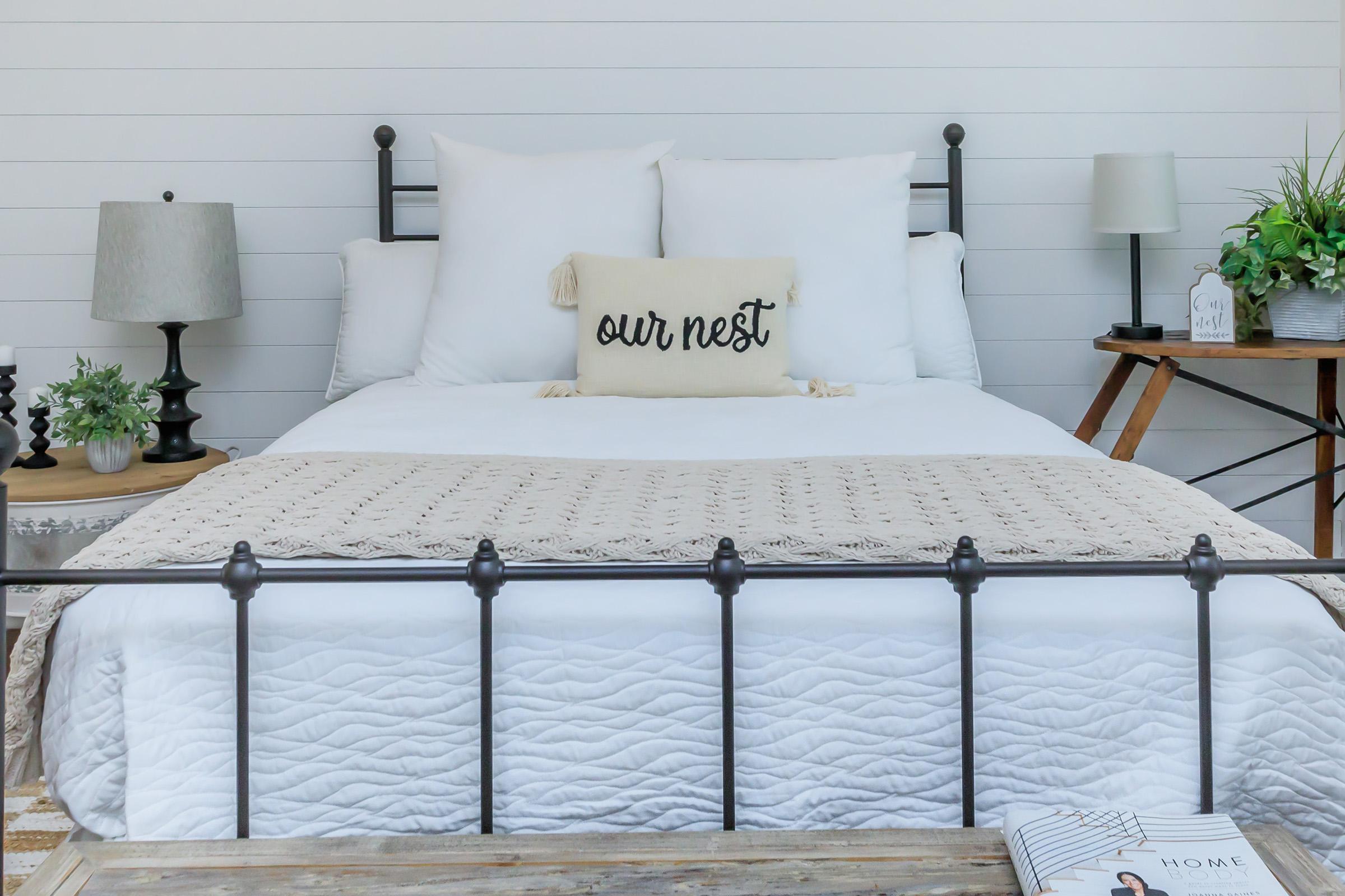 A cozy bedroom featuring a neatly made bed with white linens and a decorative pillow that reads "our nest." Flanking the bed are two bedside lamps on black stands, and a wooden side table with a plant. A light-colored knitted blanket is draped over the bed, enhancing the inviting atmosphere.