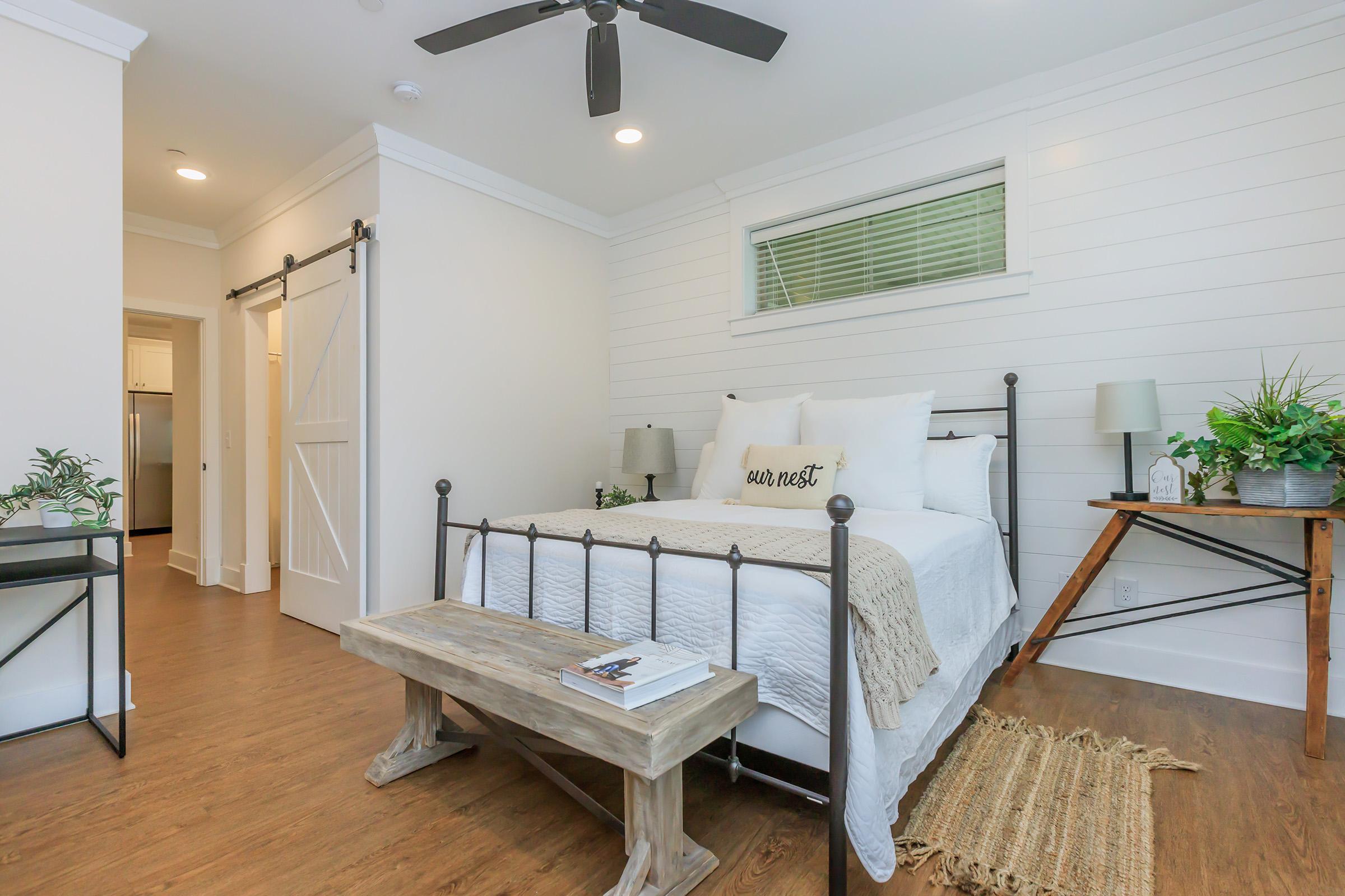 A cozy bedroom featuring a metal bed frame with white bedding and a decorative pillow. There are two bedside lamps and greenery in pots on either side. A wooden bench is at the foot of the bed, and a woven rug is on the floor. The room has a warm, inviting atmosphere with light-colored walls and natural light from a small window.