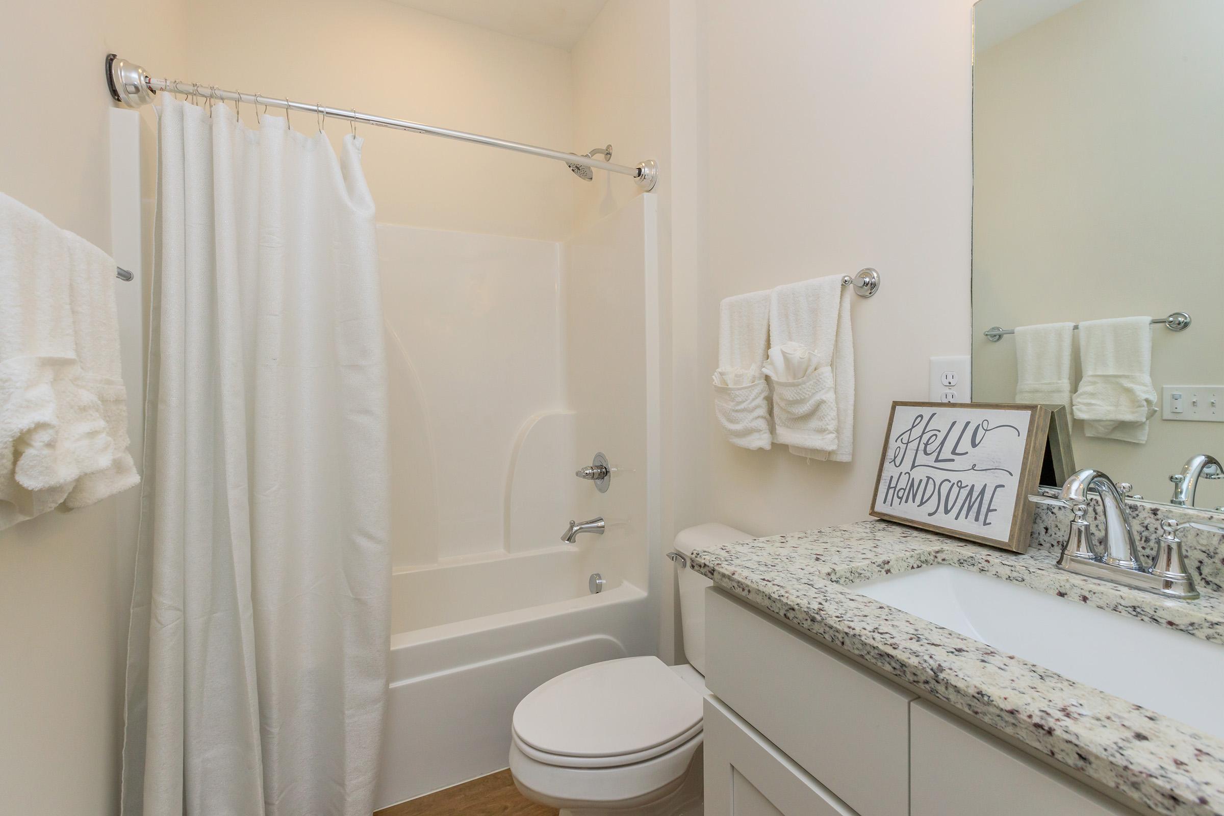 A bright and clean bathroom featuring a bathtub with a white shower curtain, a toilet, and a double vanity with a granite countertop. Soft white towels hang on the walls, and a decorative sign reading "Hello Handsome" is placed on the countertop, adding a charming touch to the space.