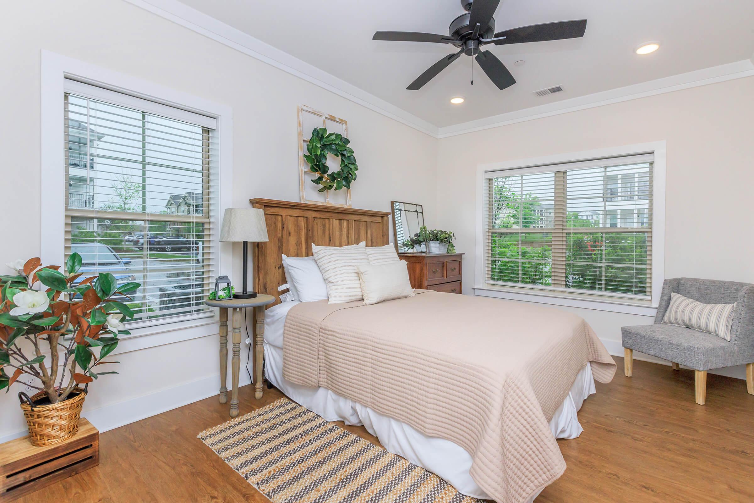 A cozy bedroom featuring a wooden bed with a beige blanket, two large windows with blinds, a small gray chair, a dresser, and a decorative plant in a woven basket. Natural light fills the room, creating a warm and inviting atmosphere.