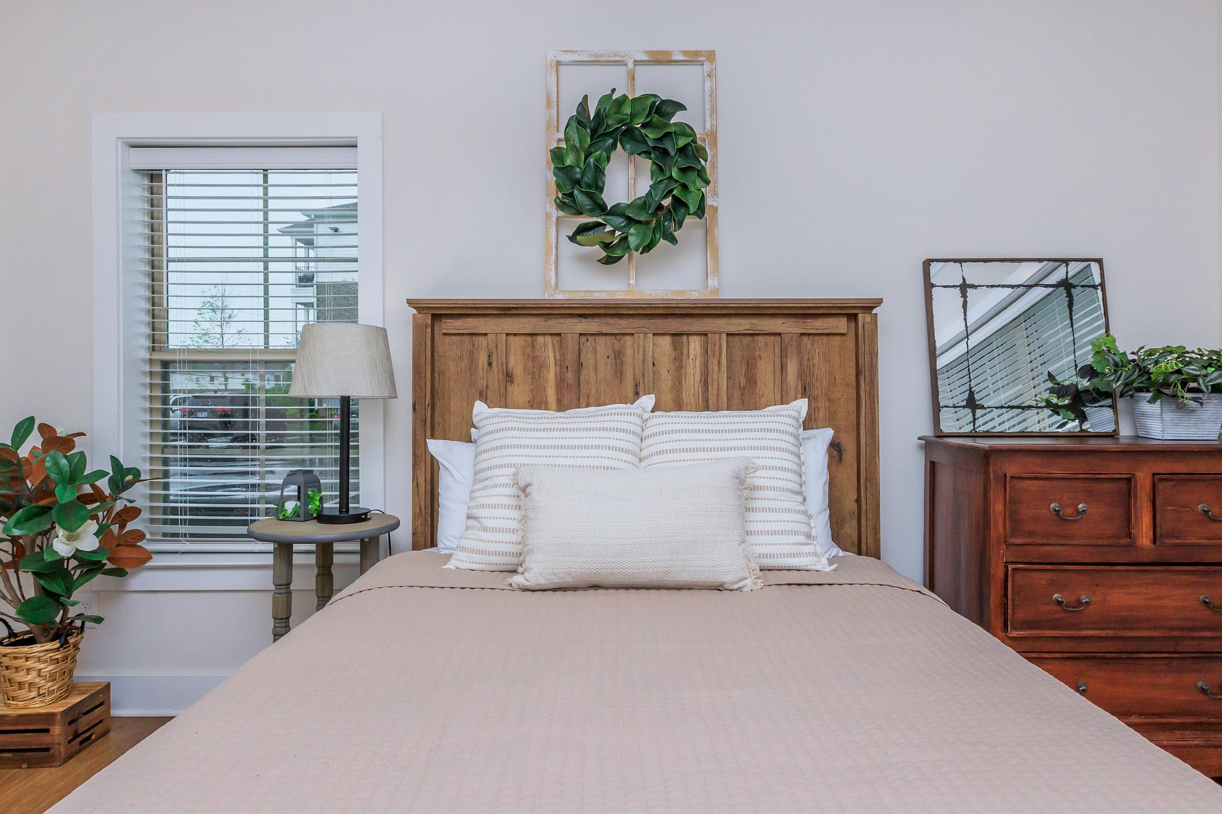 A cozy bedroom featuring a wooden headboard with decorative pillows, a bedside table with a lamp, and a mirror. A leafy wreath hangs above the bed, and a green plant sits in a basket. The room has light-colored walls and natural light coming through a window with blinds. A wooden dresser is also visible.