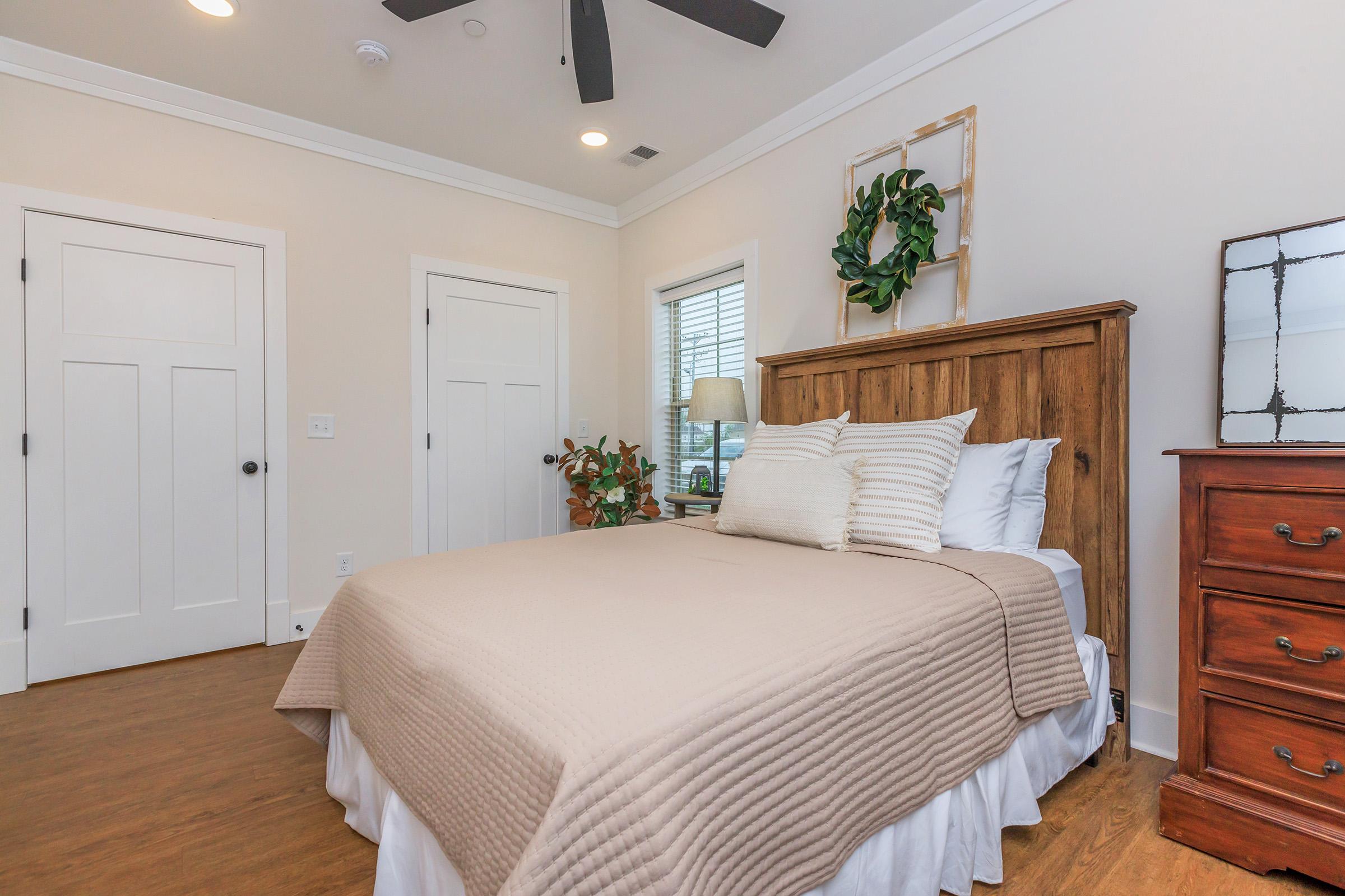 A cozy bedroom featuring a wooden bed frame with a beige quilt, two pillows, and a decorative throw. There’s a nightstand with a lamp and a small plant, along with a wooden dresser. The room has white walls, a ceiling fan, and a window with privacy blinds, creating a warm and inviting atmosphere.