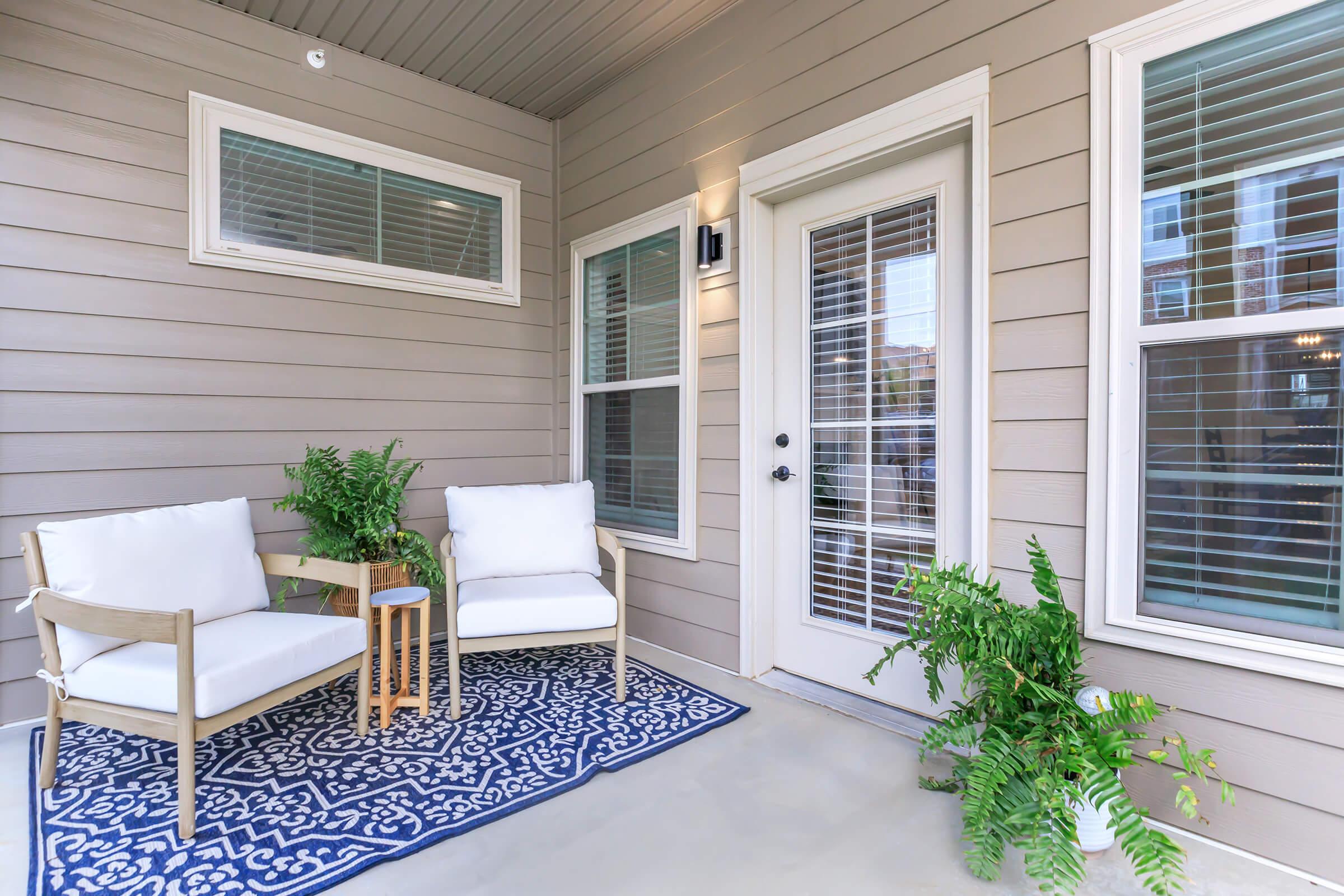 A cozy porch area featuring two white chairs and a small wooden table. There's a decorative blue and white rug on the floor, and a potted plant adds a touch of greenery. The walls are a light beige, complemented by several windows with white blinds. A door leads to the interior space.