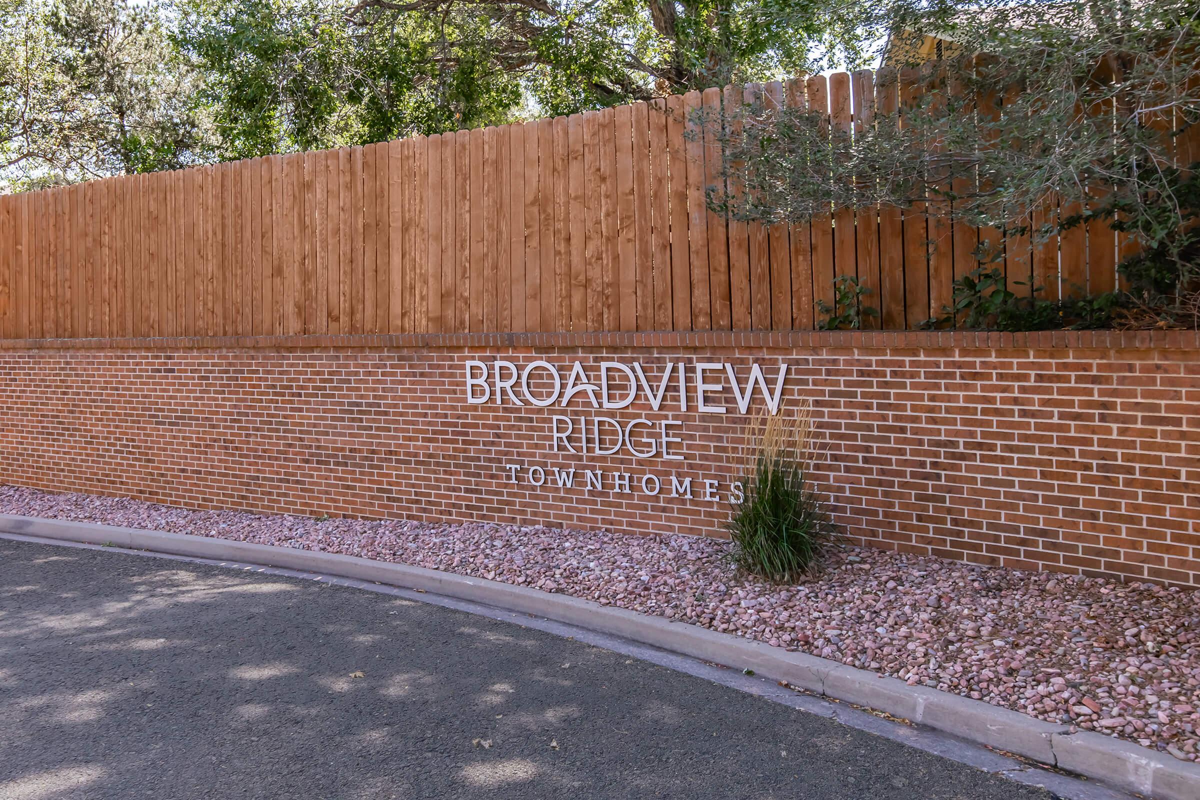 Sign for Broadview Ridge Townhomes, featuring a brick wall with white lettering. The background includes a wooden fence and some greenery, with pink pebbles along the base. The scene is set in a residential area with a clear sky and trees in the background.