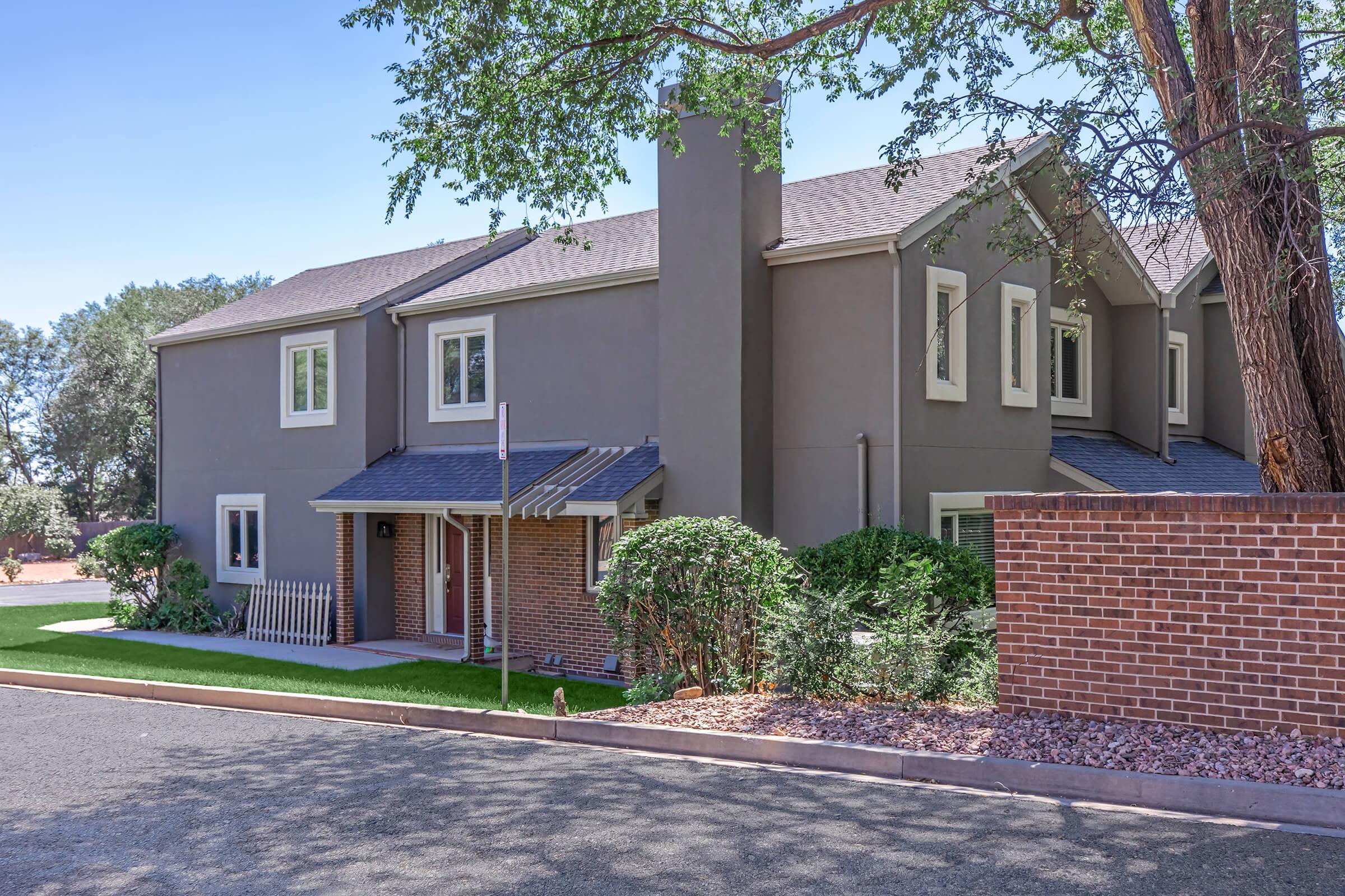 A modern two-story residential building with a gray exterior, featuring windows and a sloped roof. There are neatly trimmed bushes and a small lawn in front, along with a brick wall bordering the property. The surrounding area includes trees and a clear blue sky.