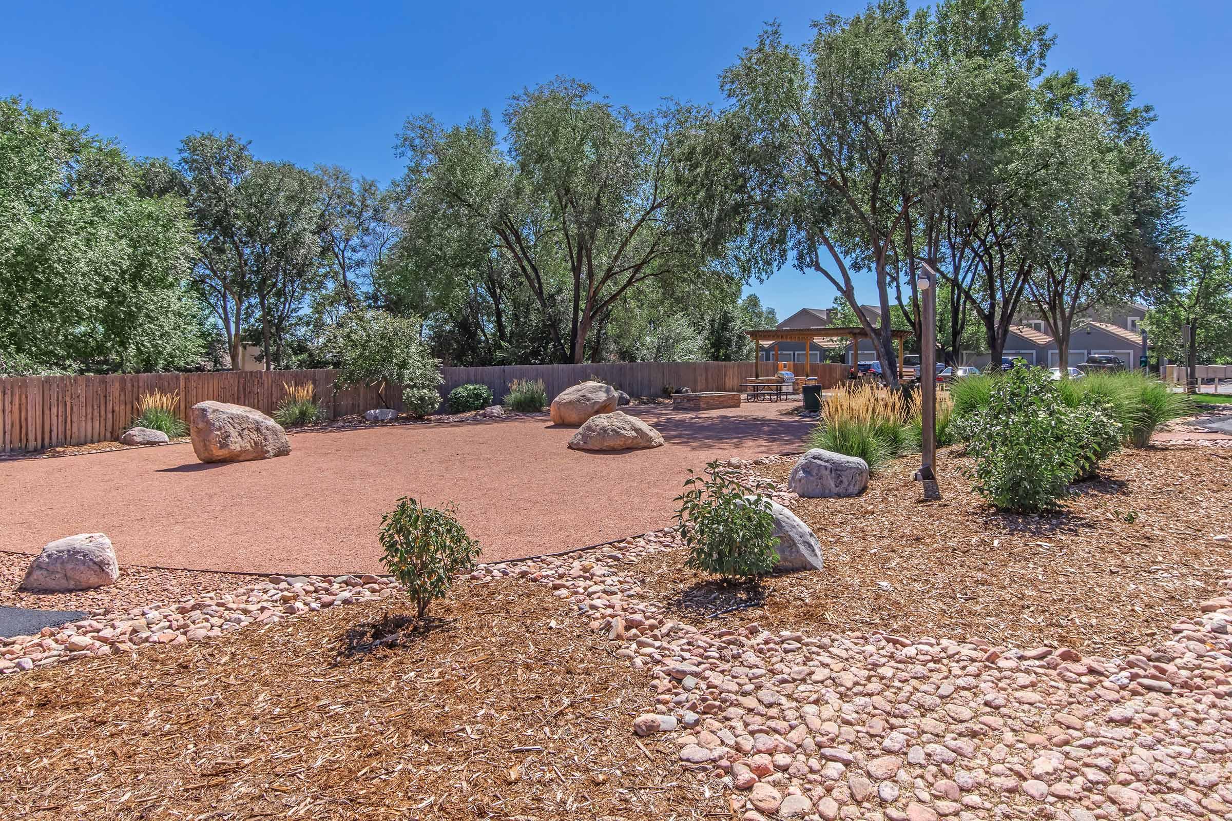 A landscaped outdoor area featuring scattered rocks and gravel, surrounded by trees. There are shrubs and a wooden fence in the background, along with a bench visible in the distance. The sky is clear and blue, indicating a sunny day.