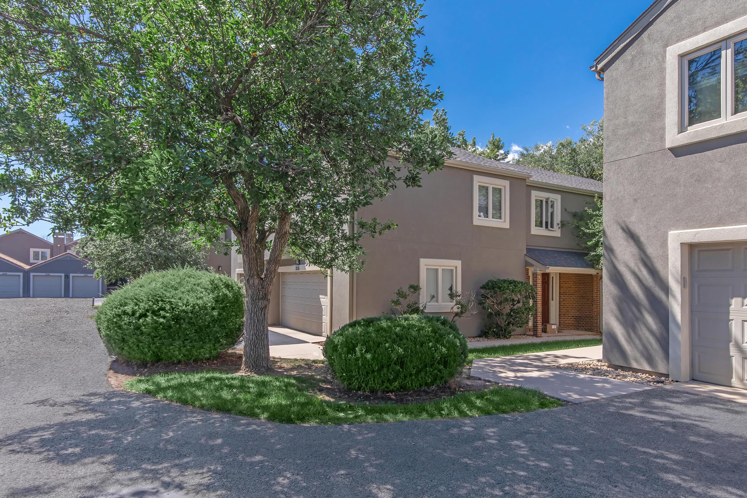 A well-maintained home with a light grey exterior, surrounded by lush green shrubs and trees. The driveway curves around the house, leading to a garage. Clear blue skies and bright sunlight highlight the inviting landscape, creating a peaceful residential atmosphere.