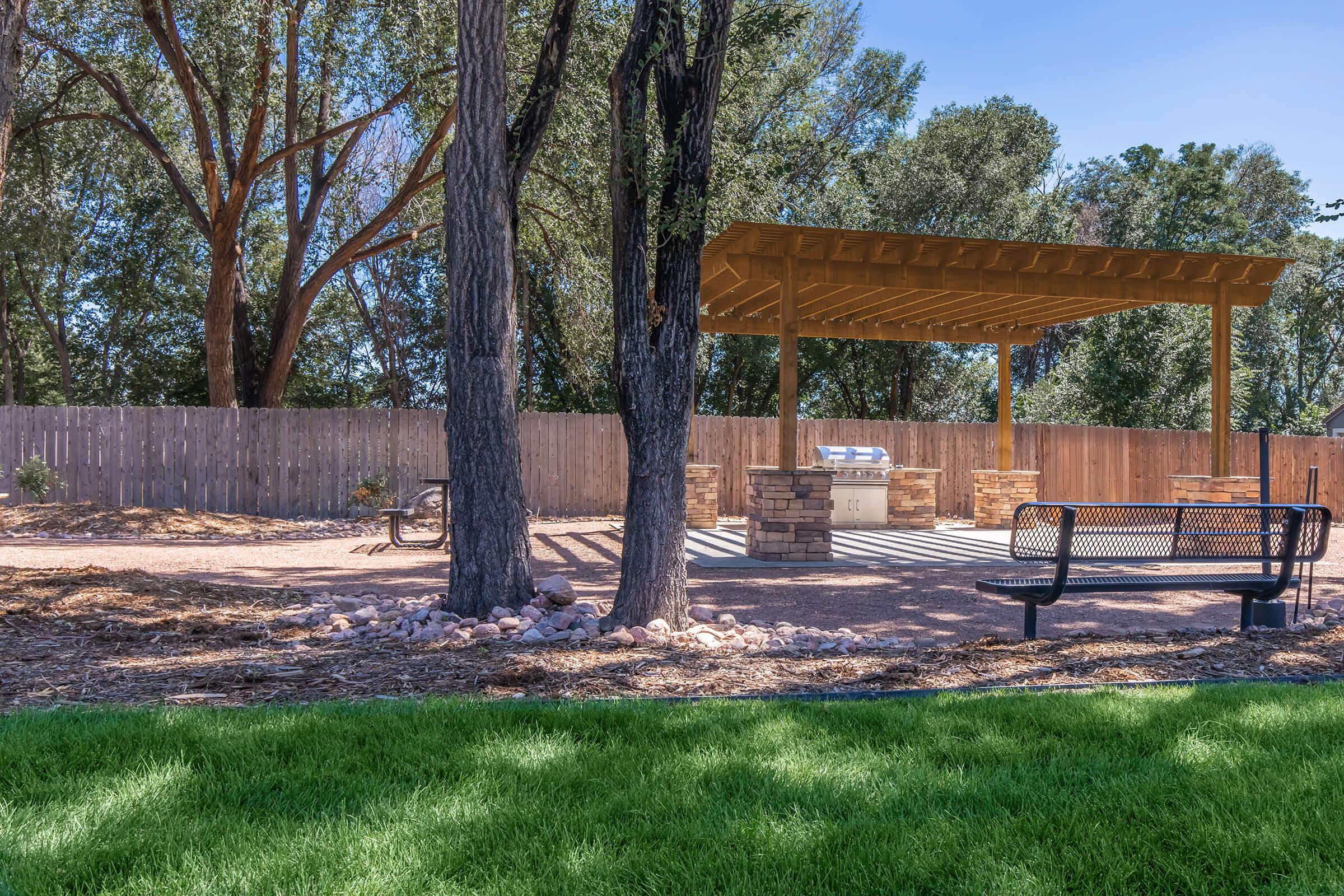 A landscaped outdoor area featuring two large trees, a shaded pavilion with stone grill stations, benches, and a grassy section. The background includes a wooden fence and a serene environment with greenery and sunlight. Perfect for gatherings and picnics.