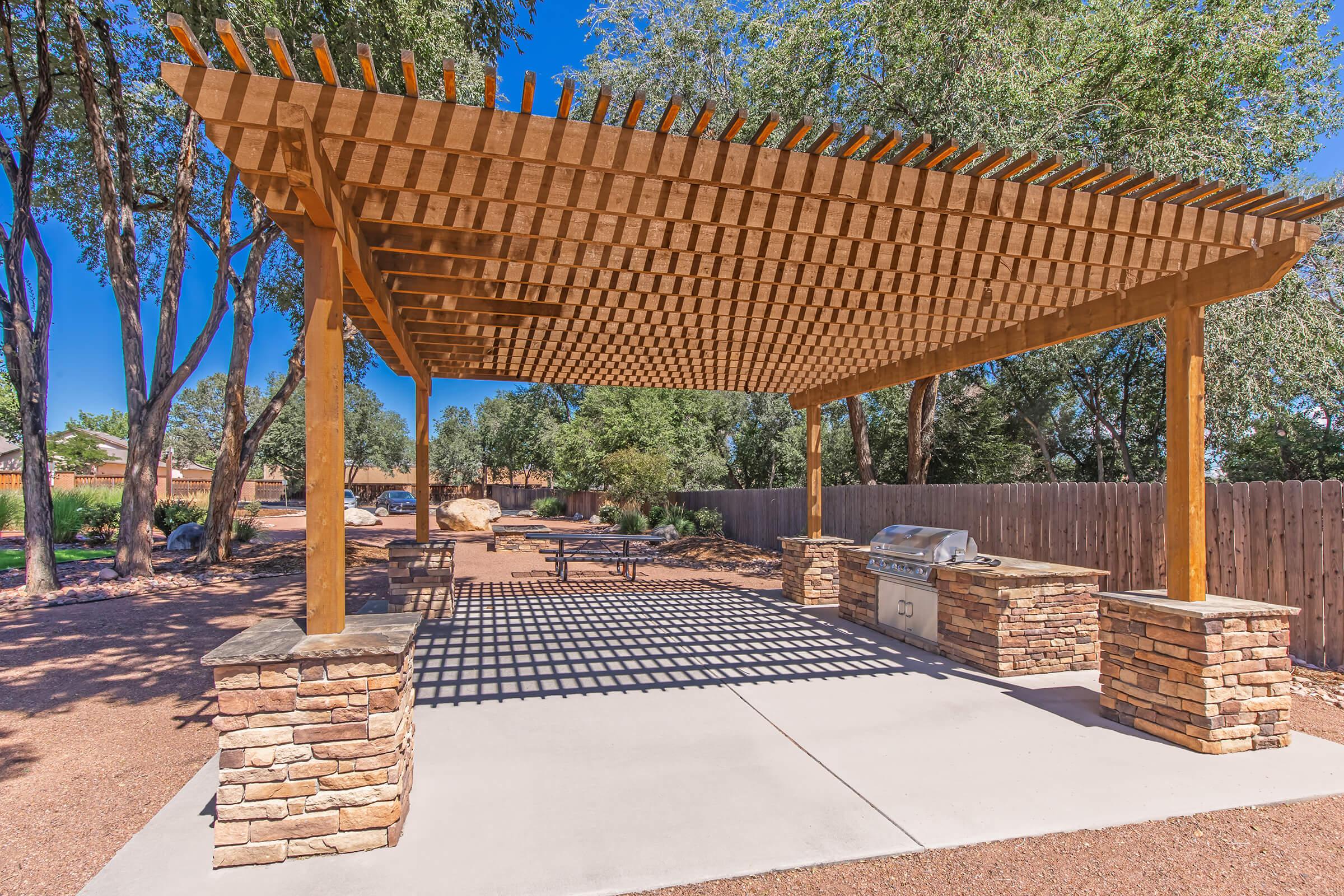 A shaded outdoor BBQ area featuring a wooden pergola supported by stone pillars, with a concrete surface and a grill. Surrounding trees provide greenery, and a stone pathway leads to the setup. The scene is bright and sunny, ideal for outdoor gatherings.