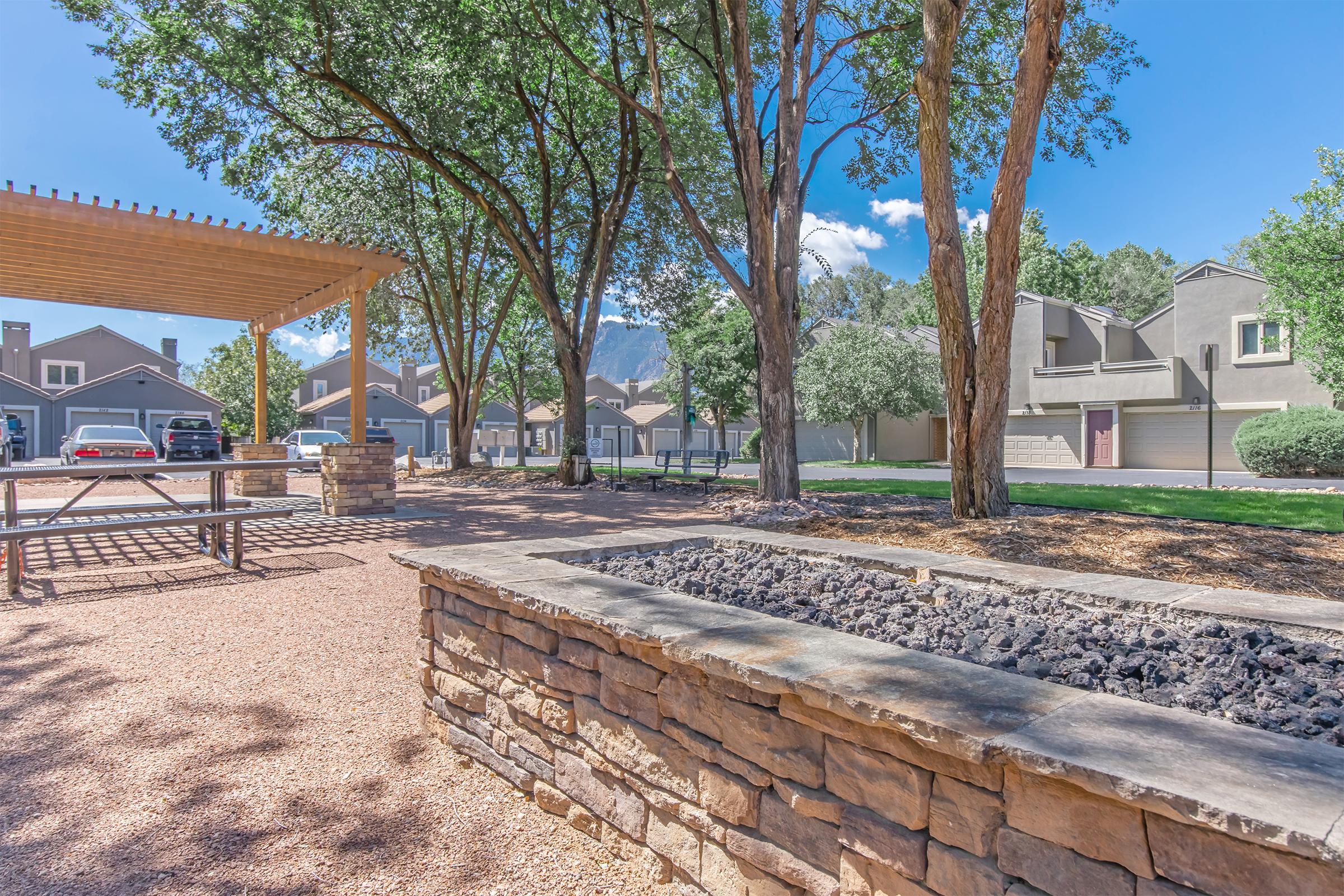 A peaceful outdoor space featuring a stone fire pit surrounded by gravel, picnic tables, and lush green trees. In the background, there are parked cars and residential buildings with a clear blue sky above, creating a serene atmosphere for relaxation and gatherings.