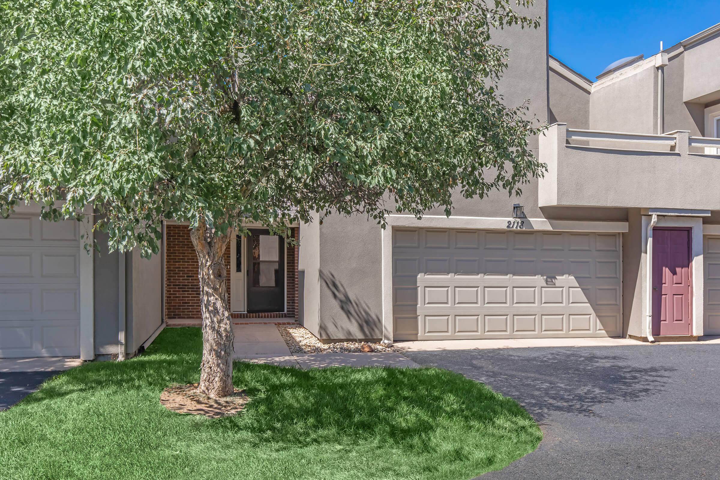 A residential exterior view featuring a gray building with a front door, a large leafy tree in the foreground, and two garage doors. The driveway is paved, and there is a patch of green grass near the tree. The sky is clear and blue, creating a bright atmosphere.