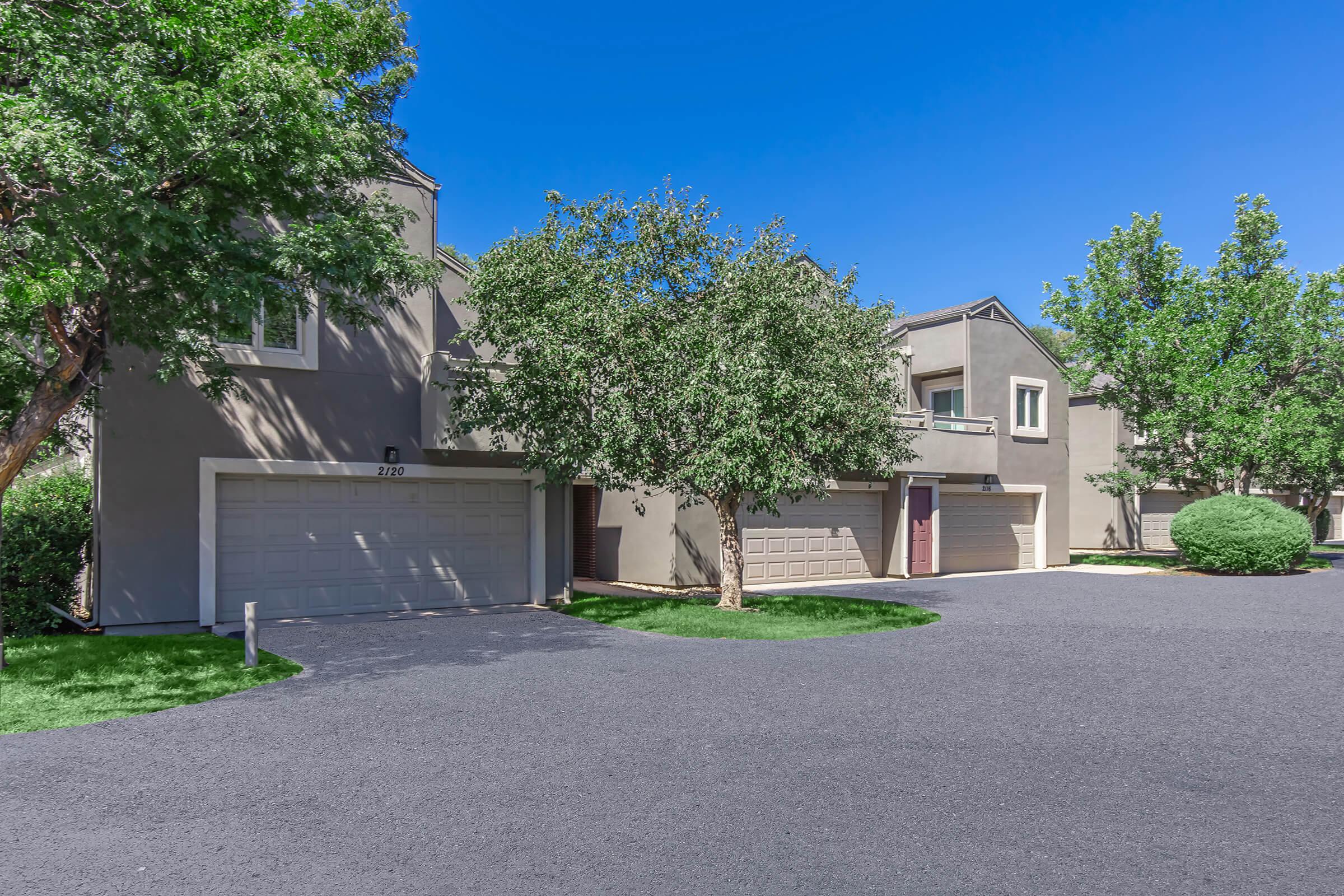 A residential street view showcasing two multi-story buildings with attached garages. The scene features well-maintained landscaping with green trees and shrubs, and a smooth dark asphalt driveway, under a clear blue sky. The overall appearance is sunny and inviting.