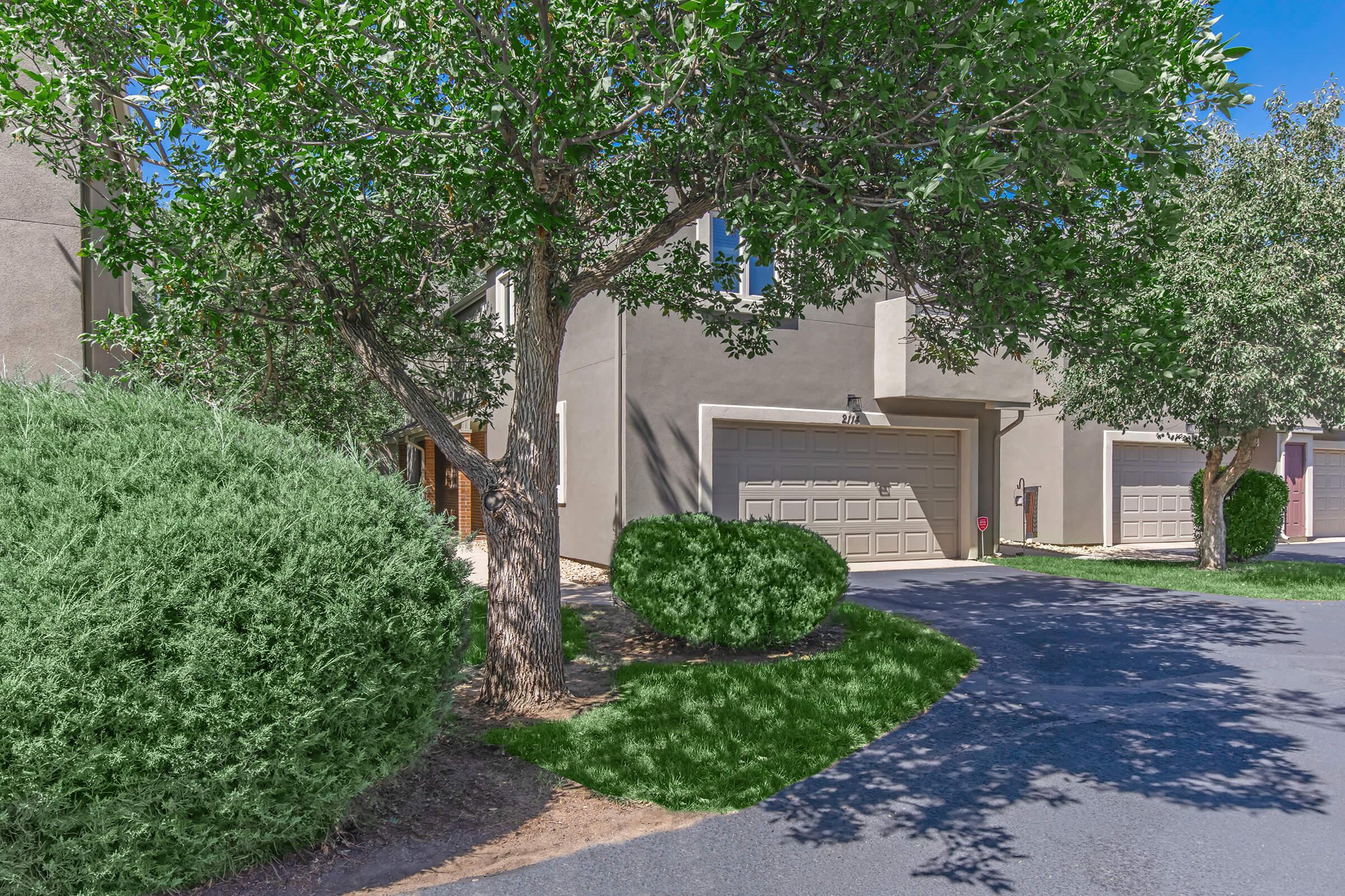 A well-maintained residential area featuring a grey house with a garage, surrounded by green shrubs and trees. A paved driveway leads to the garage, and the sky is bright blue, adding to the inviting atmosphere. The landscape is neatly trimmed, providing a tidy appearance.
