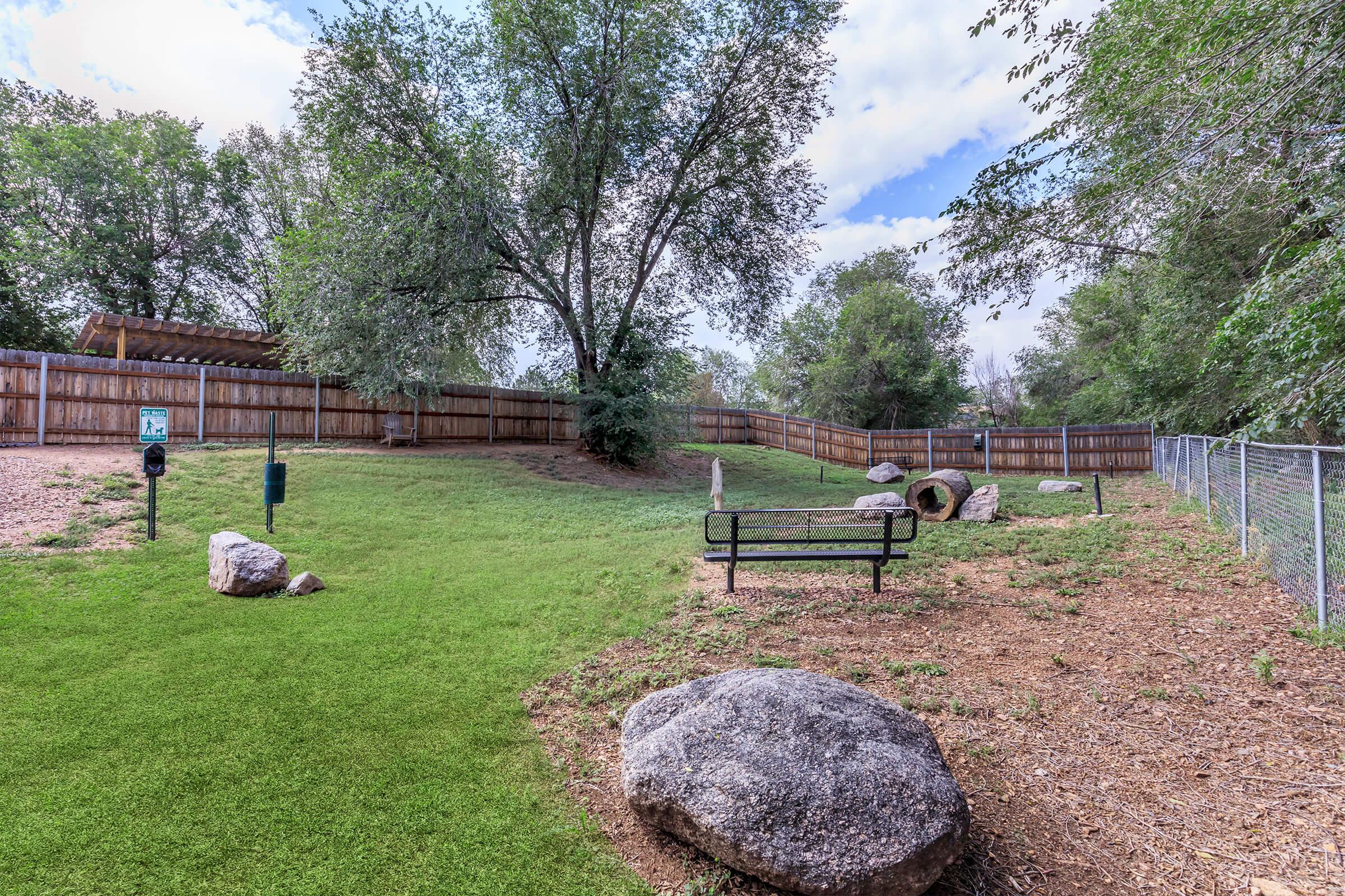 A green, grassy area in a fenced garden featuring a bench, large rocks, and trees. The sky is partly cloudy, and there's a wooden structure in the background, surrounded by a wooden fence. The overall scene is serene and inviting, perfect for outdoor relaxation.