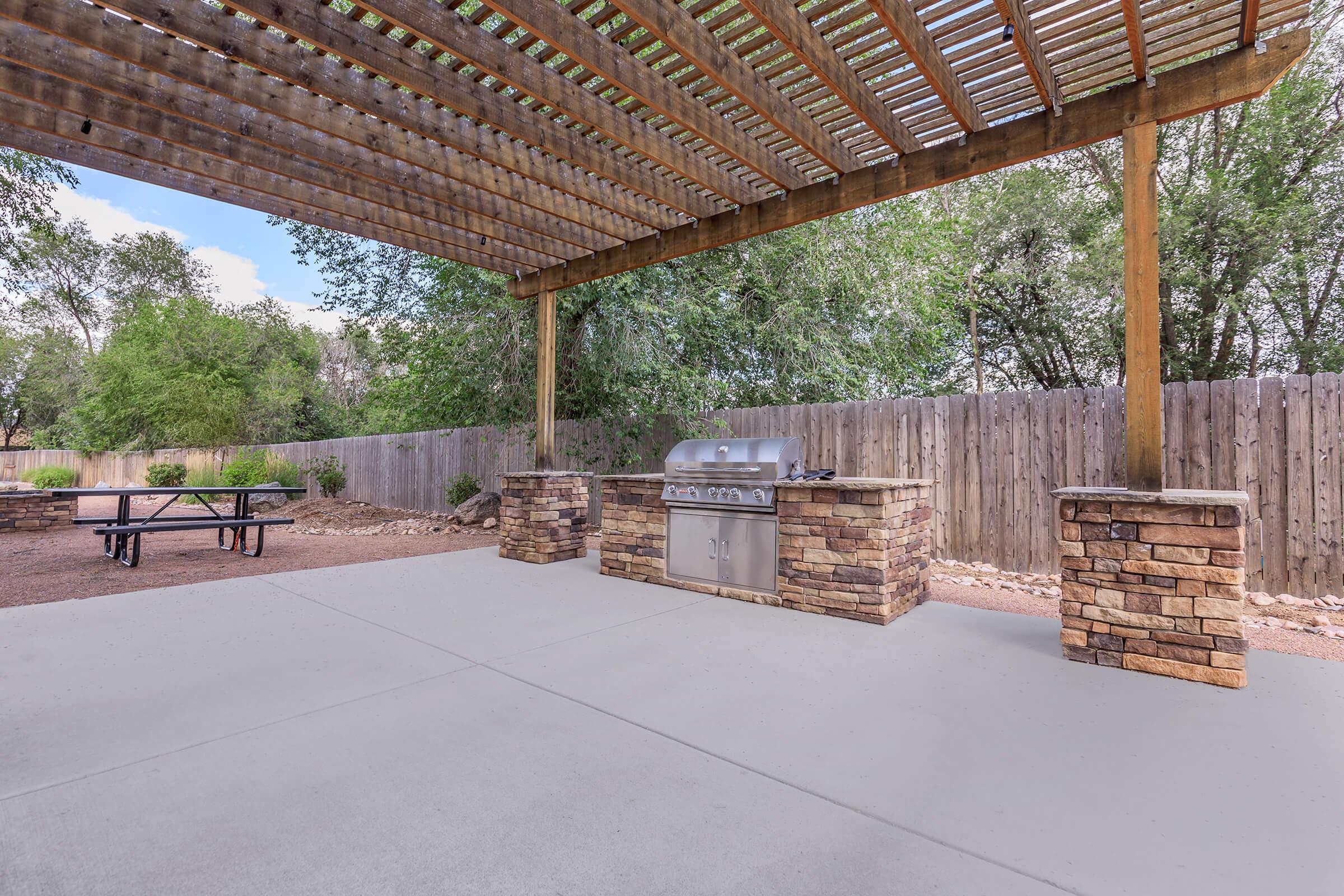 An outdoor cooking area featuring a stainless steel grill set on a stone countertop, surrounded by wooden support beams and a lush green backdrop. The area includes a concrete floor and a picnic table in the distance, nestled under a pergola structure.
