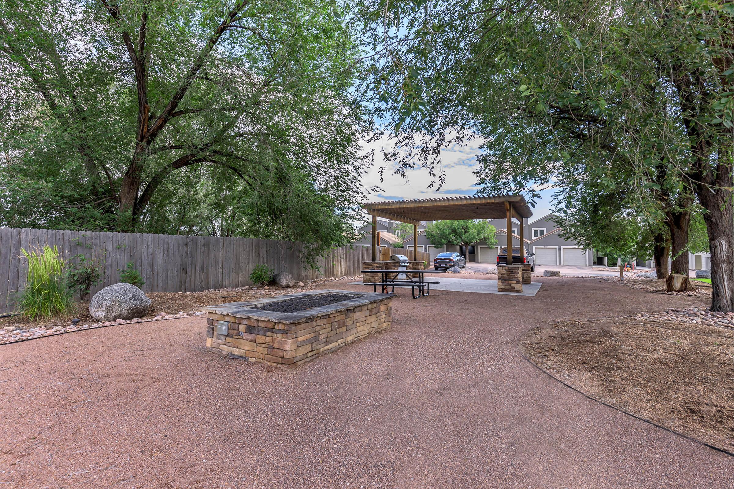 A landscaped outdoor area featuring a gravel path, a stone fire pit, and a shaded pavilion with seating. Surrounding trees provide shade, and a wooden fence encloses the space. In the background, there are multiple garages visible, adding to the functional outdoor setting.