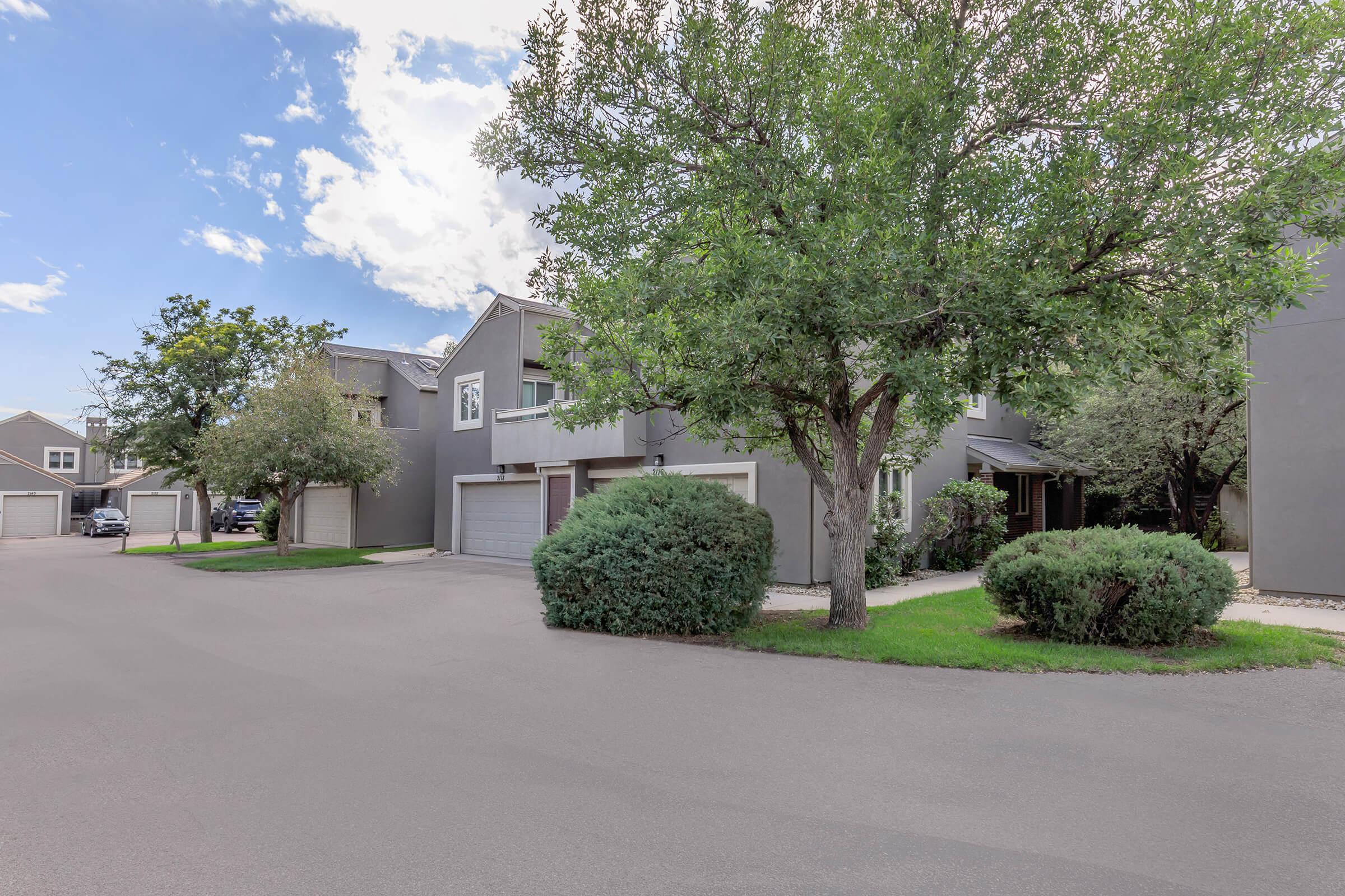A peaceful residential street featuring gray townhouses with attached garages, surrounded by lush green trees and shrubs. The sky is partly cloudy, creating a serene atmosphere. The road is well-maintained, leading into the neighborhood.