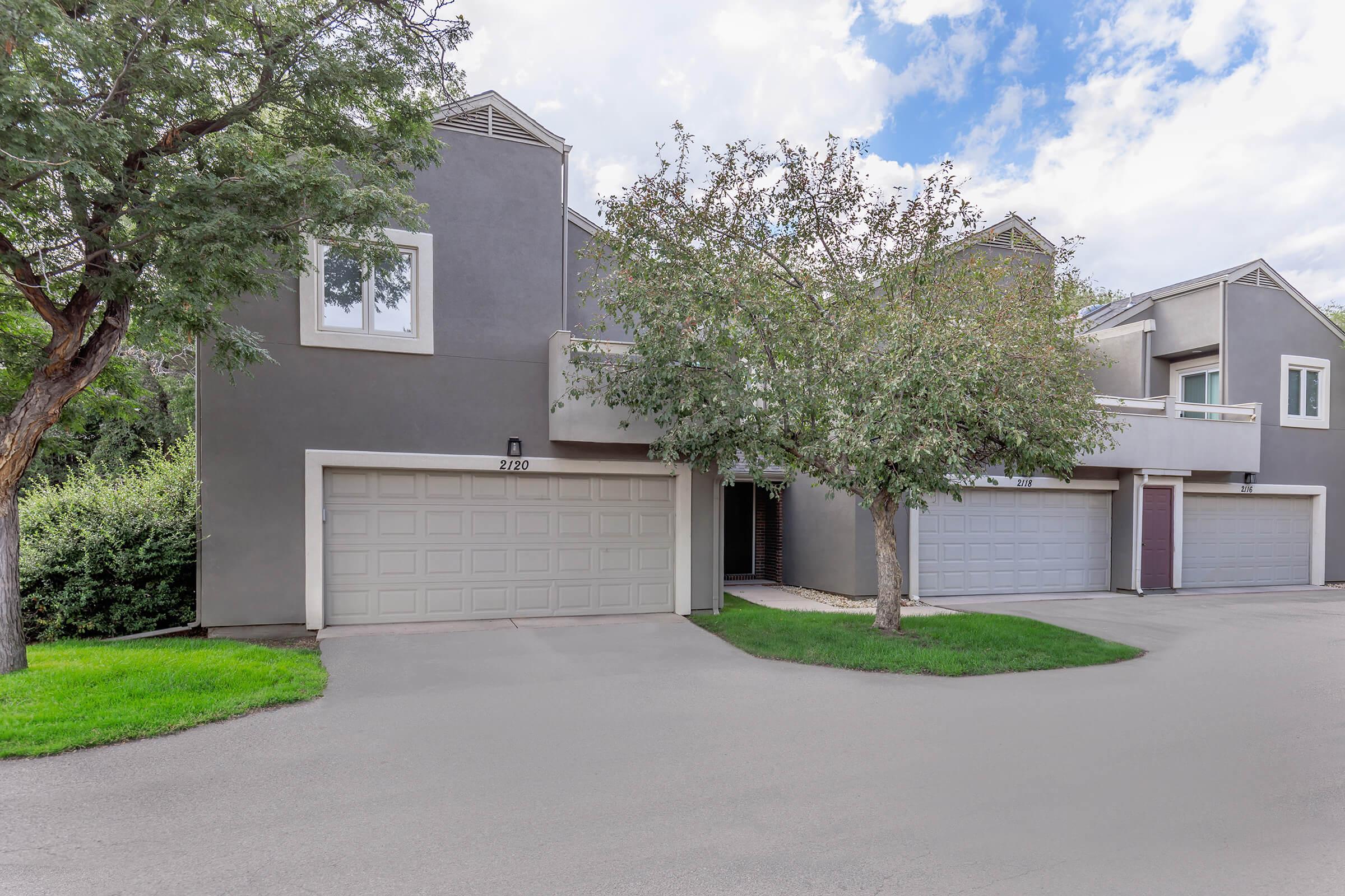 A modern, gray multi-unit residential building with garage doors and a landscaped front yard. The structure features large windows and is surrounded by trees and greenery under a partly cloudy sky. The scene conveys a peaceful suburban setting.