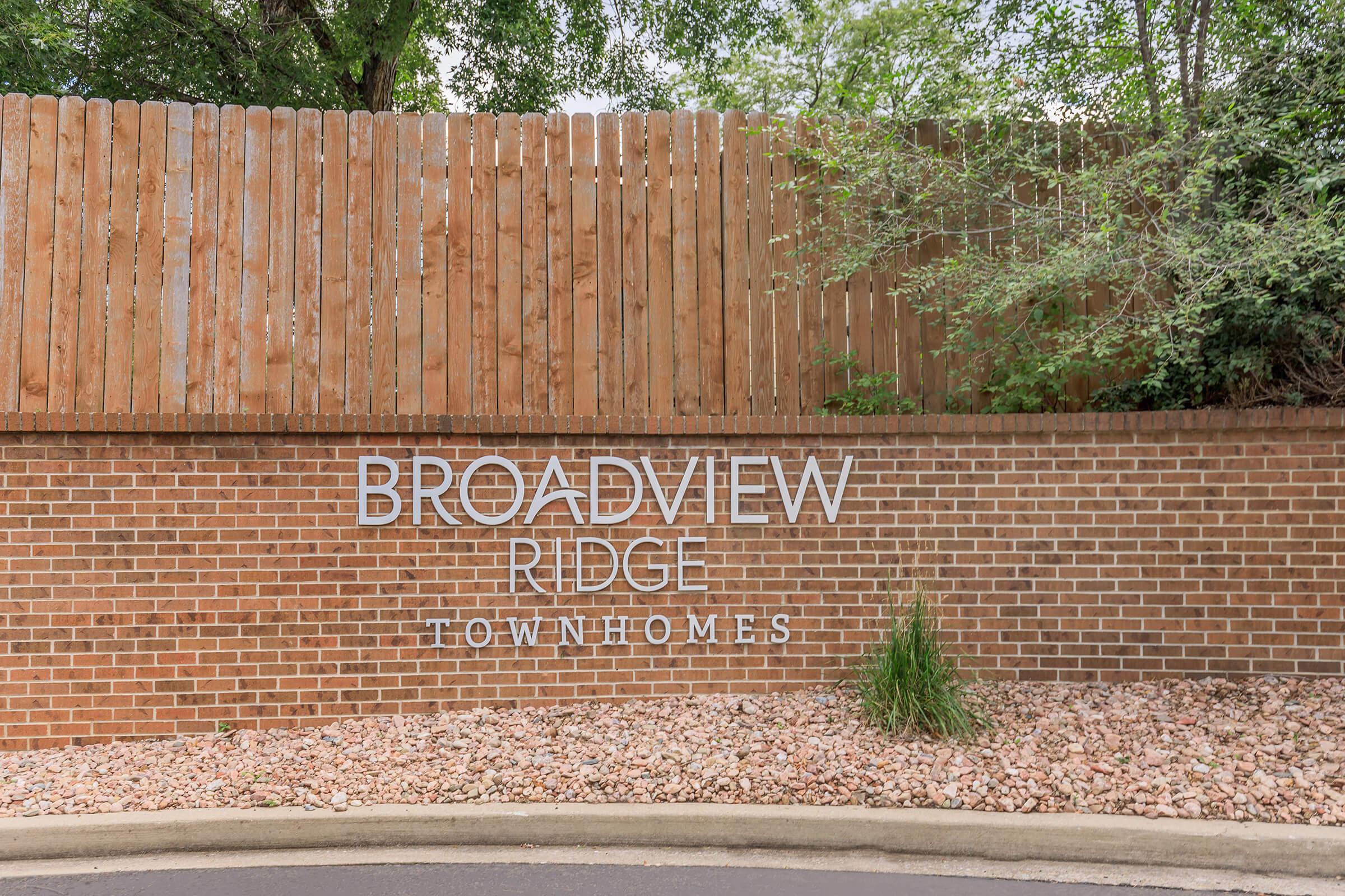 Signage for Broadview Ridge Townhomes, featuring modern lettering on a brick wall with a landscaped area in front. The backdrop includes a wooden fence and greenery, creating a welcoming atmosphere.