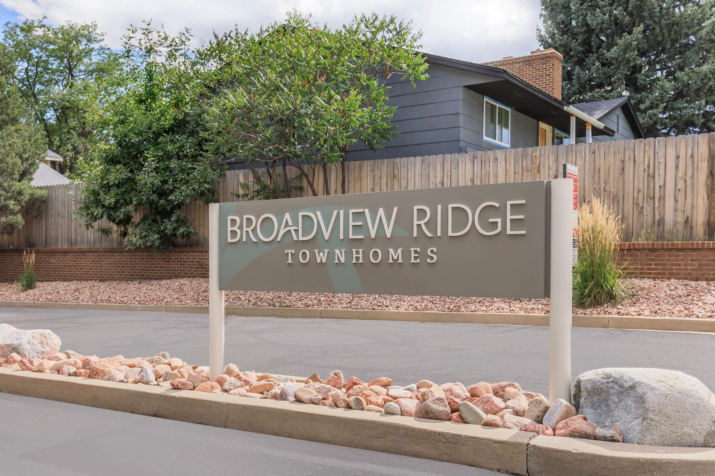 Sign for Broadview Ridge Townhomes, featuring the community name prominently displayed on a modern sign. The background includes a residential building, trees, and landscaping with rocks along the base. The sky is mostly clear with some clouds.