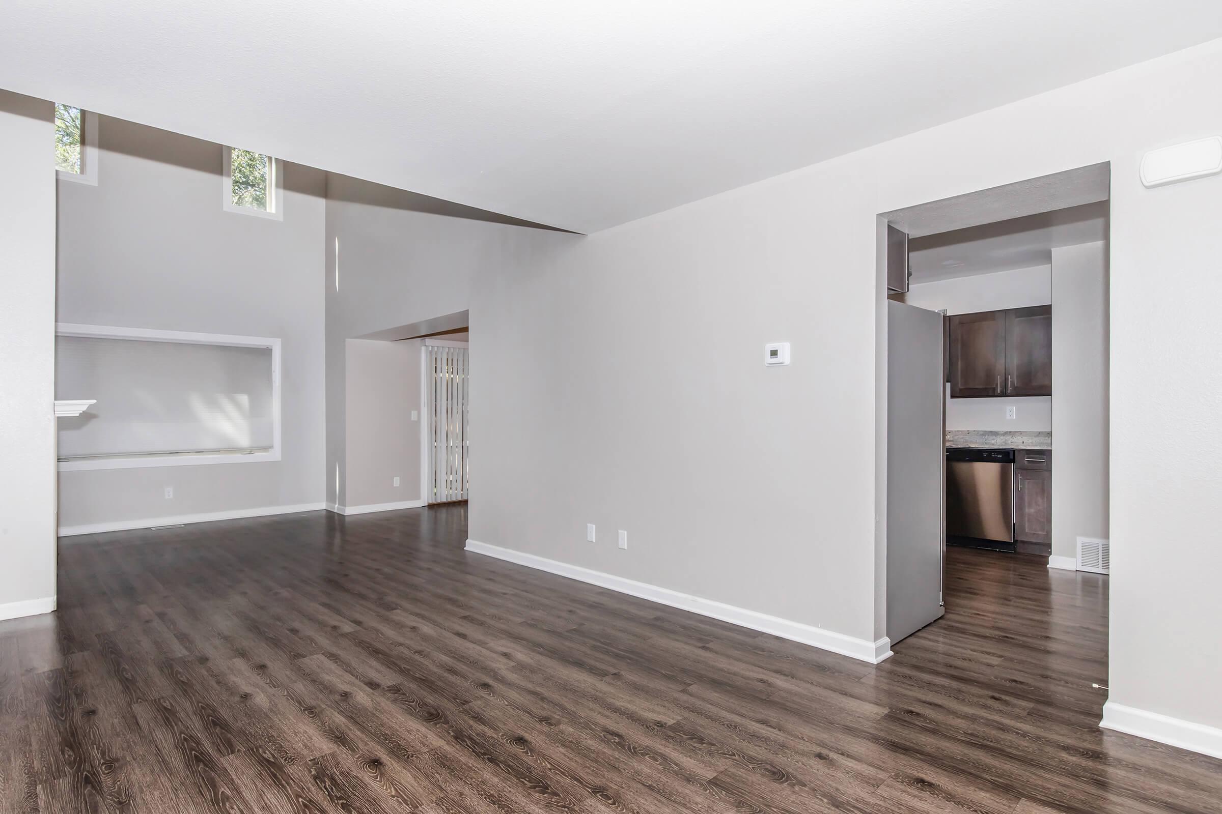 Spacious interior of a modern living room with high ceilings, wooden flooring, and neutral-colored walls. A doorway leads to a kitchen area, while large windows allow natural light to fill the space. The room features an open layout, perfect for various furniture arrangements.