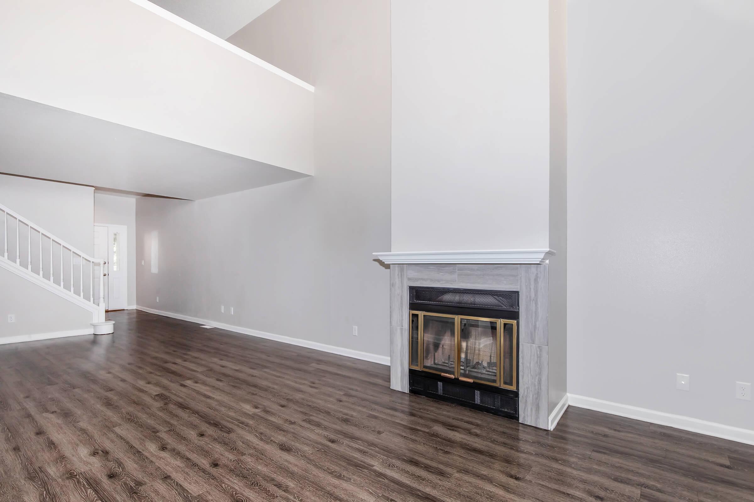 A spacious, modern living room featuring a large fireplace with a marble surround, hardwood flooring, and light gray walls. The room has high ceilings and a staircase visible in the background, creating an open and airy feel. Natural light floods in, enhancing the inviting atmosphere.