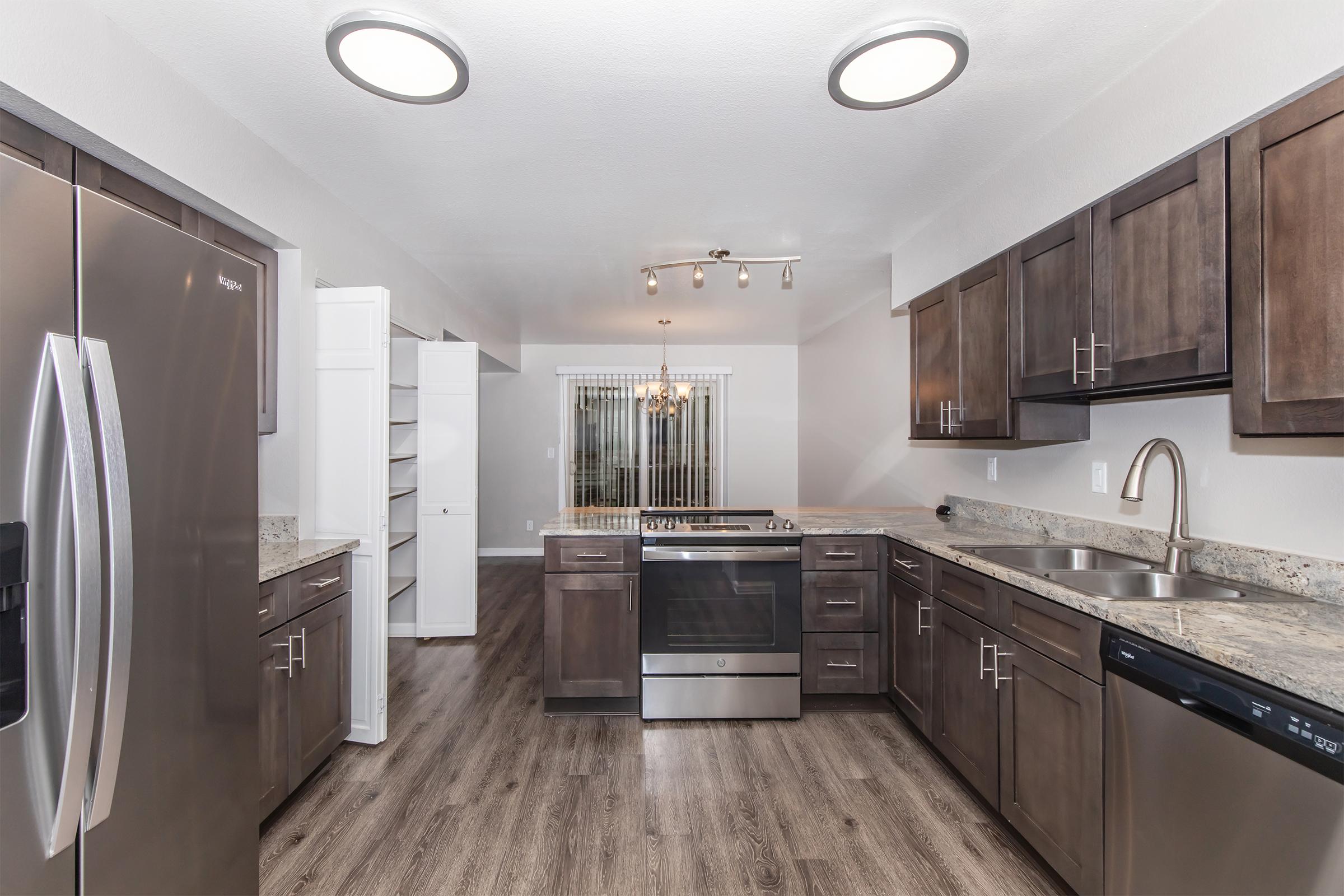 A modern kitchen featuring dark wood cabinets, stainless steel appliances including a refrigerator and stove, and a granite countertop. The kitchen has sleek lighting fixtures and an adjacent pantry. Large windows provide natural light, illuminating the spacious layout with wood-style flooring.