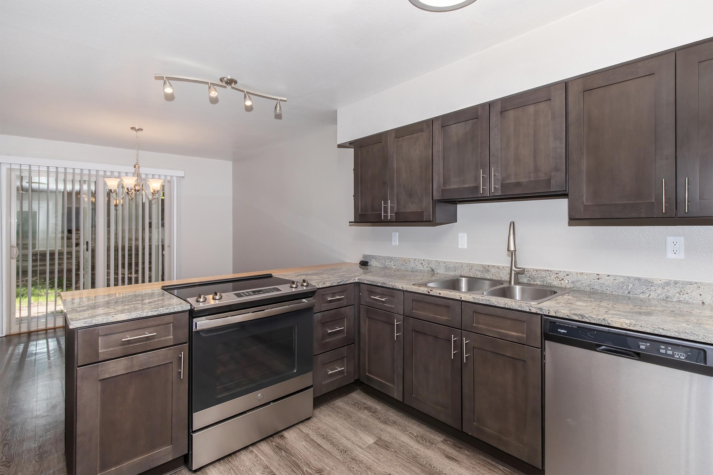 Modern kitchen featuring dark wood cabinets, stainless steel appliances, a granite countertop, and an island with a stovetop. Natural light streams in from a nearby sliding door leading to an outdoor area. A dining area with a chandelier is visible in the background.
