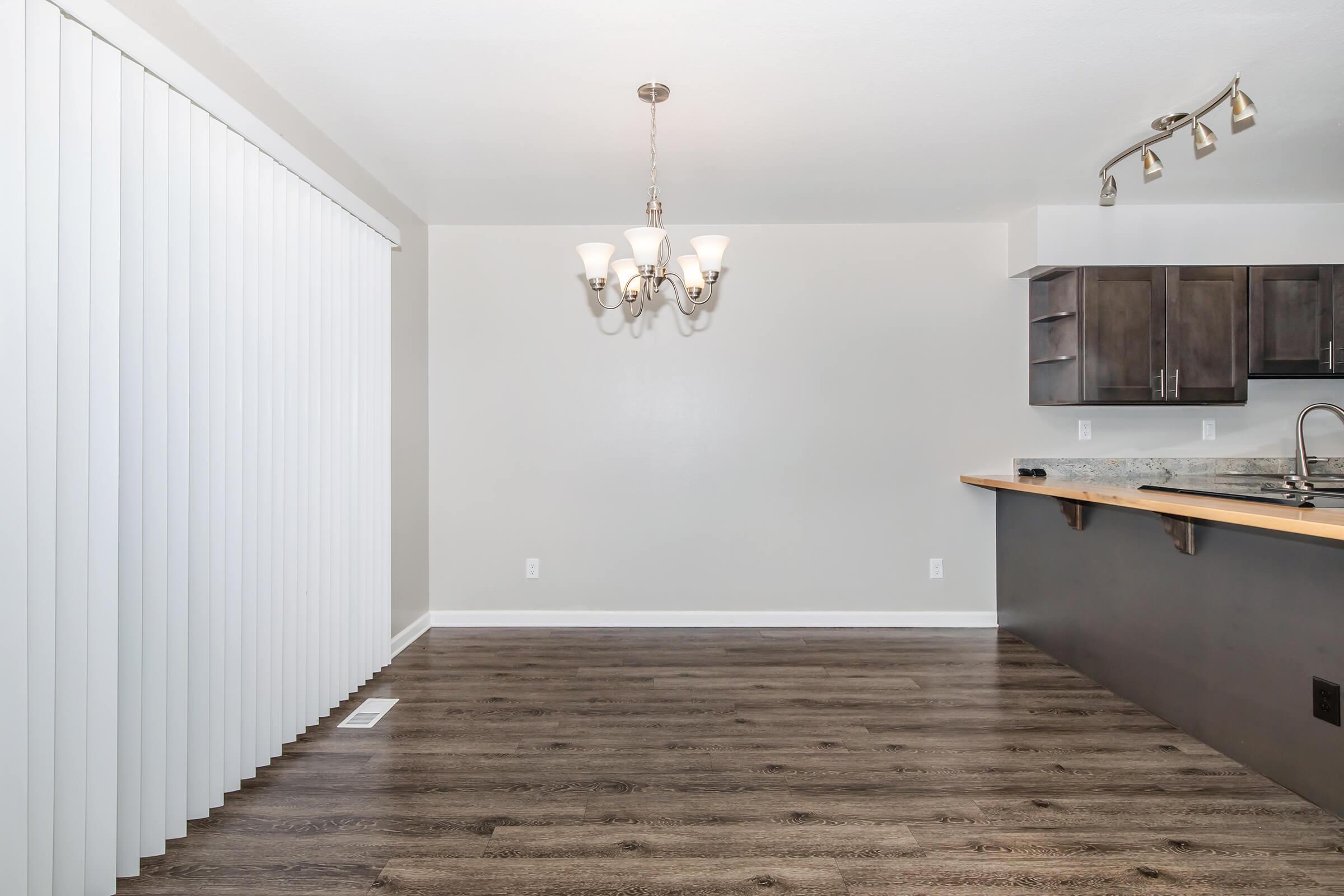 Spacious interior with light-colored walls and dark wood flooring. A dining area features a decorative chandelier overhead, while the kitchen area includes dark cabinets and a countertop. Vertical blinds cover a window, creating an open and airy atmosphere.
