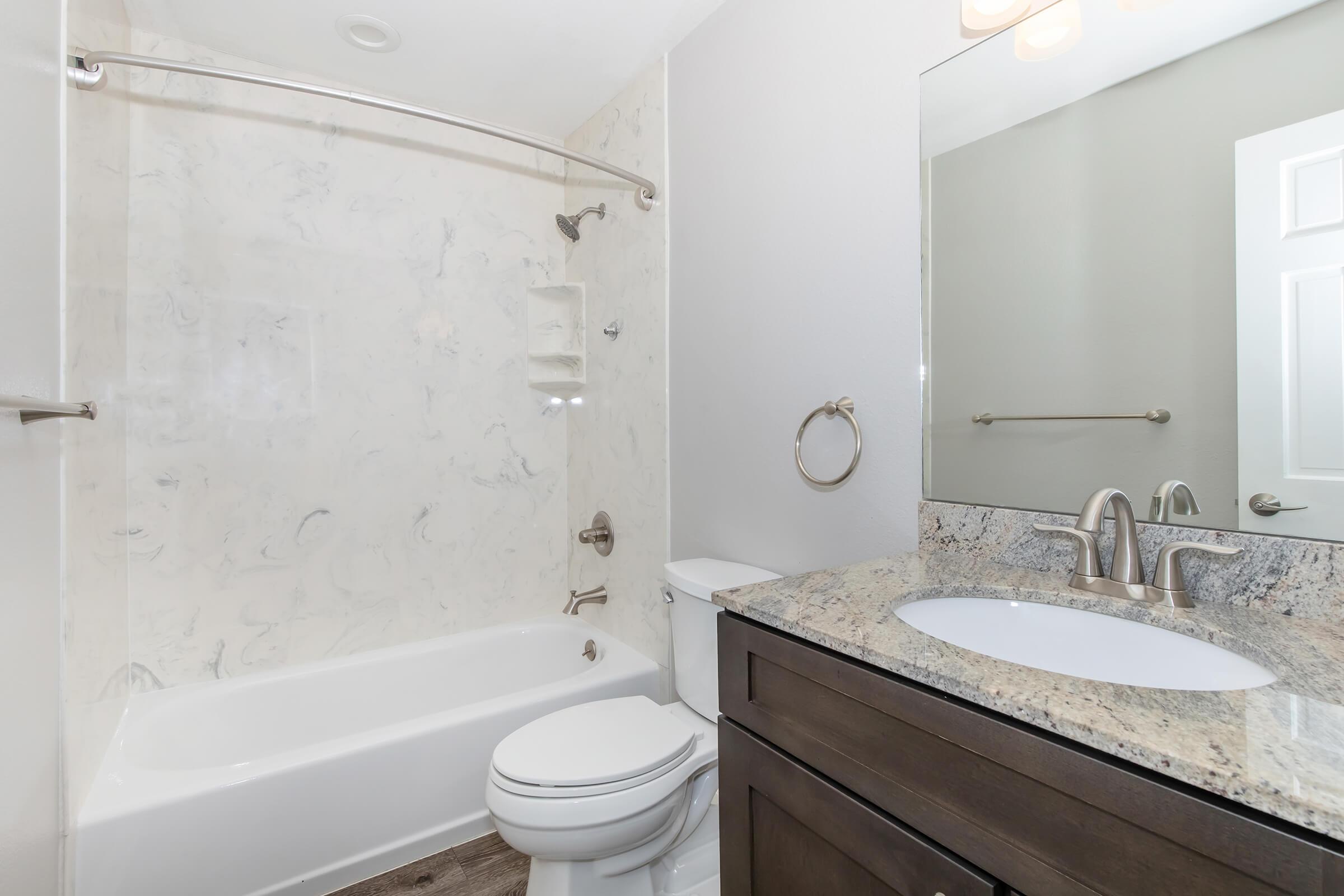 A modern bathroom featuring a white bathtub with a marble-patterned shower wall, a granite countertop on a dark wooden vanity, a circular mirror, and a small towel ring. The floor is a light wood-style material, and the space has neutral-colored walls.