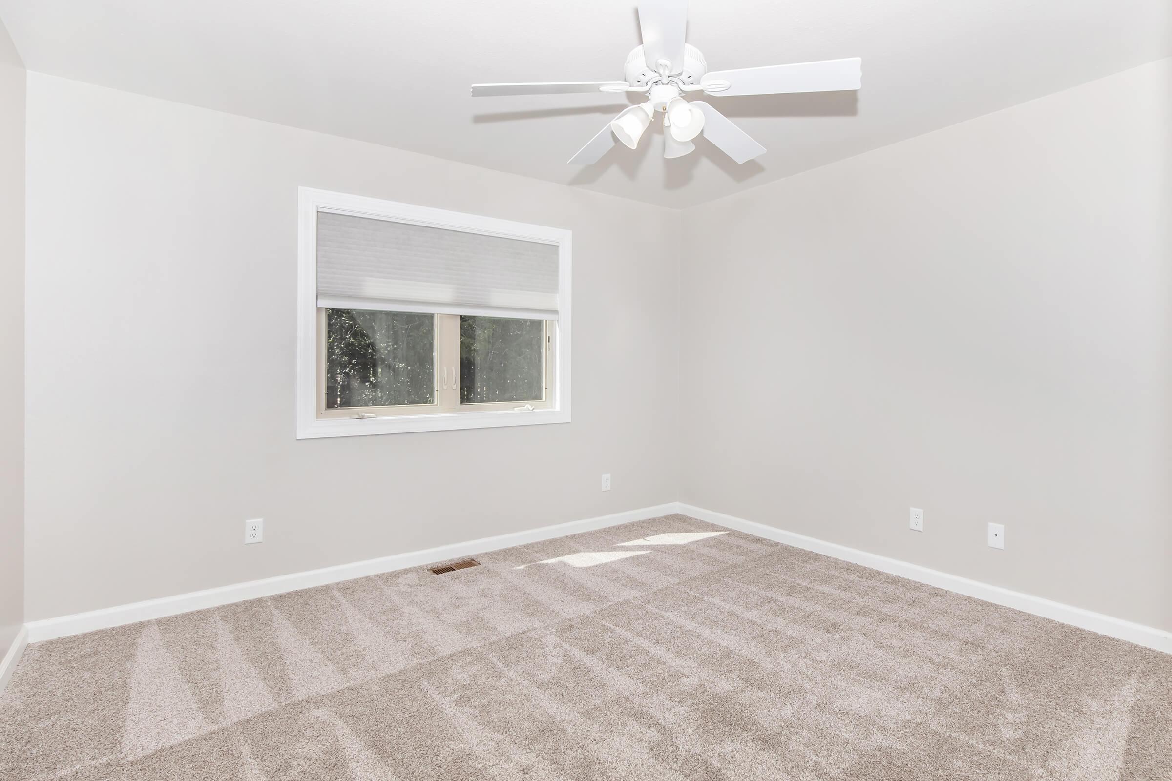 A spacious, empty room featuring light-colored walls and plush carpet. There is a ceiling fan with light fixtures and a single window covered by a light blind, allowing natural light to enter. The floor shows shadows from the window, creating a bright and airy atmosphere.