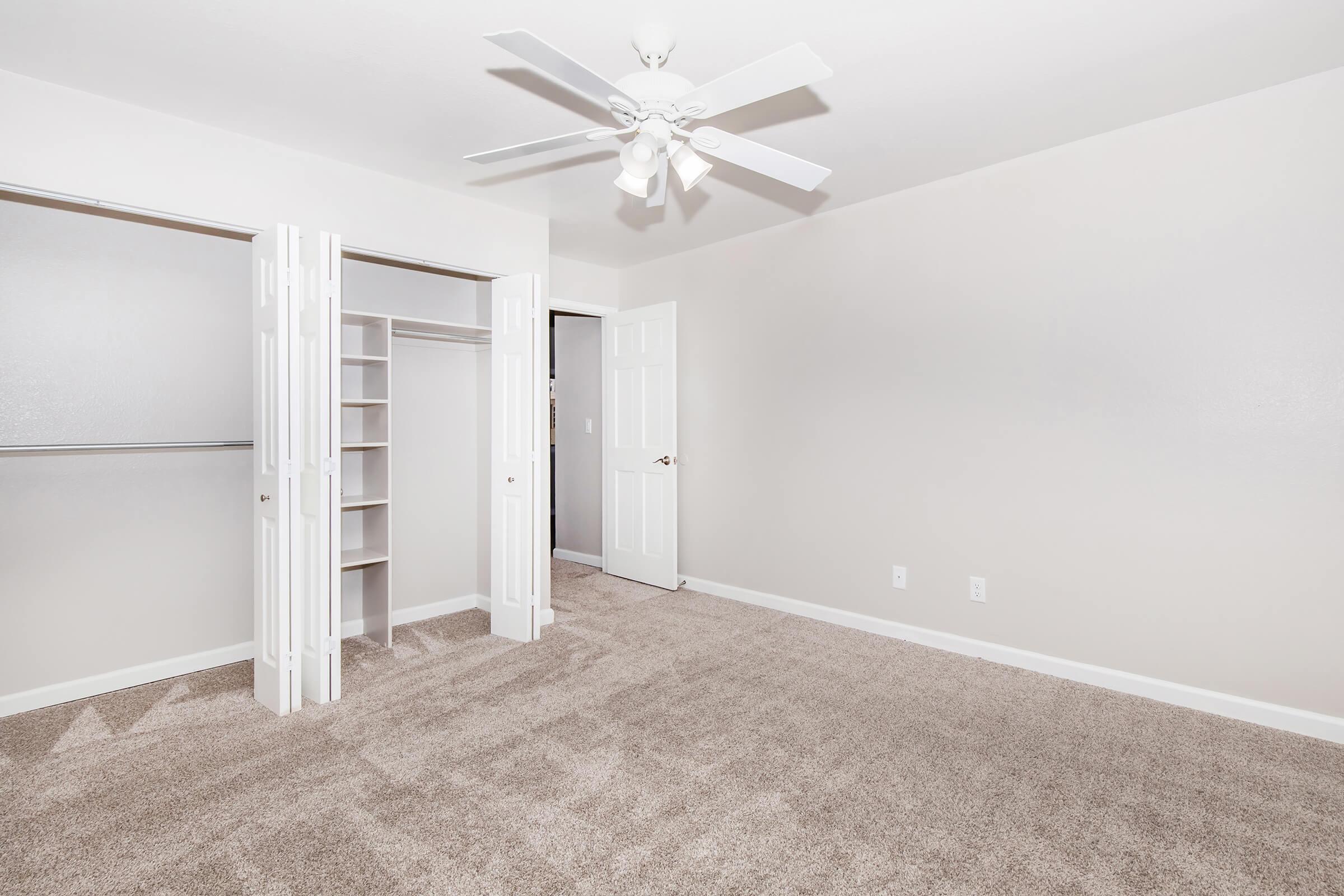 Empty room with light beige walls and carpeting. A ceiling fan is visible in the center. On one side, there is a closet with sliding doors that open to reveal shelves and hanging space. A door to another room is slightly ajar in the background, indicating the room's layout. Soft, natural light illuminates the space.