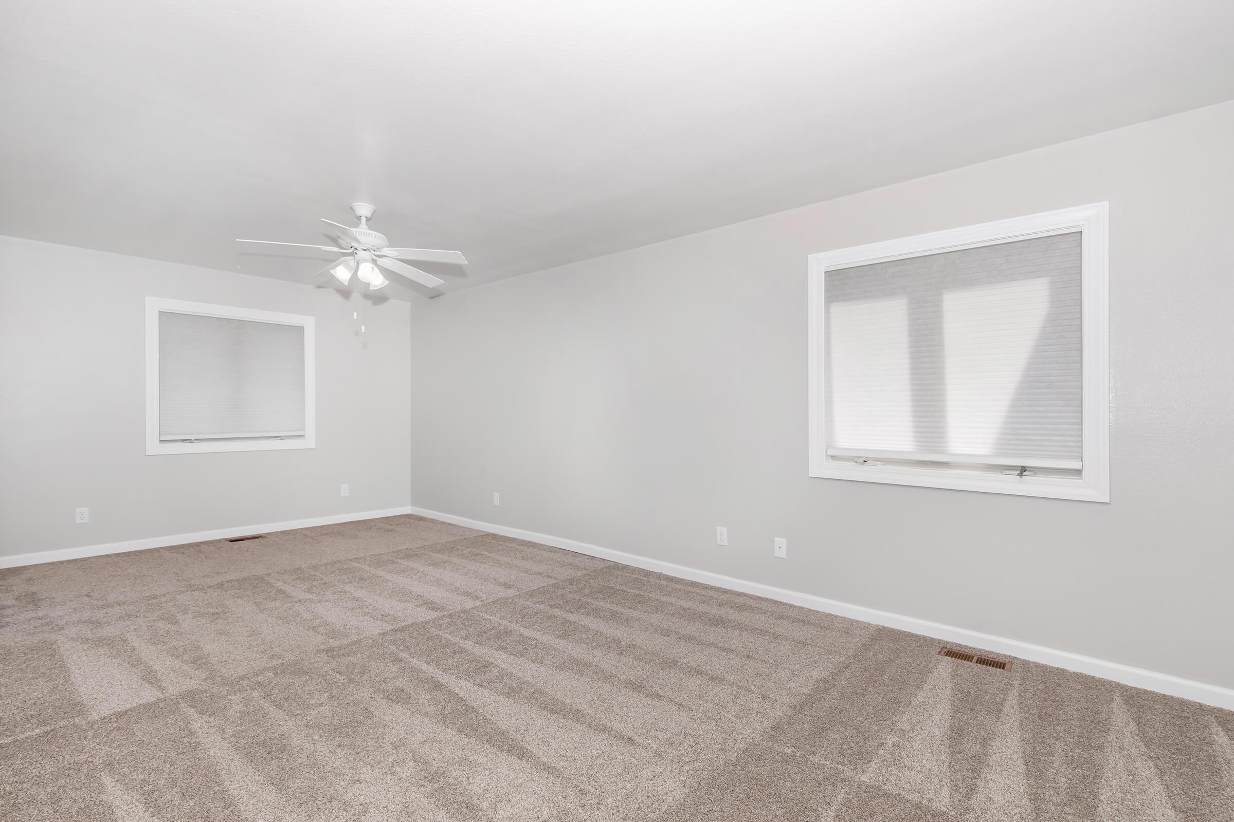 A spacious, empty living room with light gray walls and beige carpet. Two windows with blinds provide natural light. A ceiling fan is centrally mounted, and the room is devoid of furniture, creating a clean and open space.